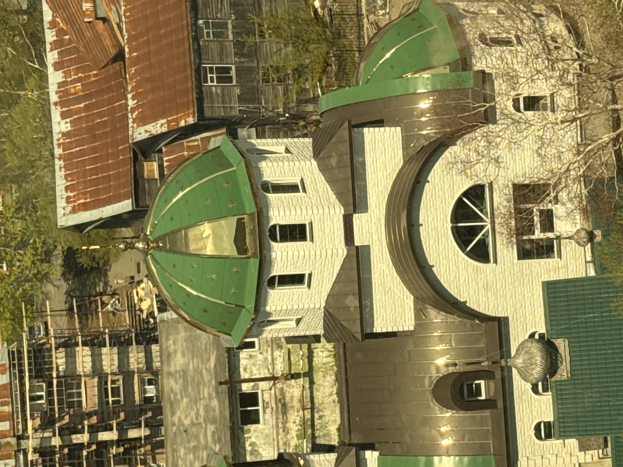 An aerial view captures a mixed urban landscape in Petropavlovsk-Kamchatsky, Russia, dominated by a prominent multi-domed structure resembling an Orthodox church. The central and largest dome is green with gilded sections and small golden star appliqués, topped with a golden finial. The church building features textured, light-colored walls, possibly stone or stucco, with various arched and rectangular windows. Portions of the roof are covered in dark, metallic material, reflecting sunlight in several spots.
To the left and slightly behind the church, a multi-story building is visible under construction or renovation, extensively covered in scaffolding. Its windows are discernible through the framework. Further to the left and closer to the foreground, a low-rise, light-colored building with a flat roof and rectangular windows is present.
To the right and behind the main church structure, several older, weathered wooden buildings with steeply pitched, heavily rusted corrugated metal roofs are observed. These structures have small windows. Sparse, bare trees, characteristic of early spring or late autumn, are scattered throughout the scene, particularly visible to the right and in the background. Green foliage is also present in the distant background. The vantage point is elevated, looking down upon the collection of buildings. No individuals are visible in the scene.
