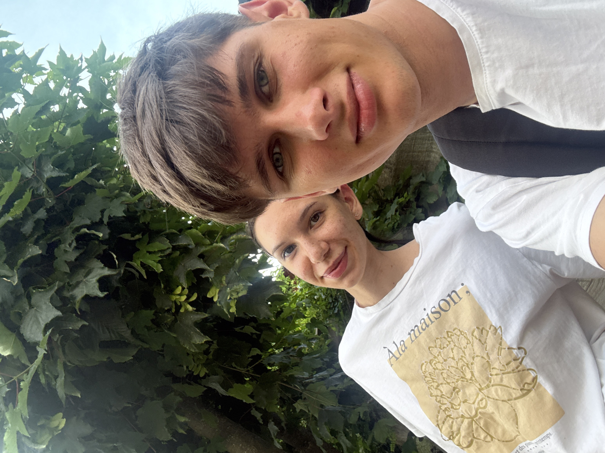 In this selfie, two young people are captured in an outdoor setting, framed by lush green foliage and a bright sky. The image is oriented such that the subjects are at an angle, with the sky and leaves forming a backdrop.
The person in the foreground, who appears to be a young man, is looking directly at the camera with a slight smile. He has short, brown hair and clear, light-colored eyes. He is wearing a white t-shirt.
Behind and slightly to the side of him, a young woman is also visible, smiling warmly at the camera. She has long, dark hair pulled back, and she is also wearing a white t-shirt. Her t-shirt features a graphic print with text. The visible text on her shirt reads "Ala maison;" followed by a flower illustration and more text below that appears to be in French.
The scene suggests a casual, daytime encounter, possibly during pleasant weather given the brightness and the presence of dense greenery. The focus is on the individuals and their cheerful expressions, indicating a happy or relaxed moment shared between them. The foliage hints at a natural environment, perhaps a park or garden. There are no explicit landmarks that identify Grindelwald, Switzerland, but the lush greenery is consistent with the region. The overall mood is lighthearted and friendly.