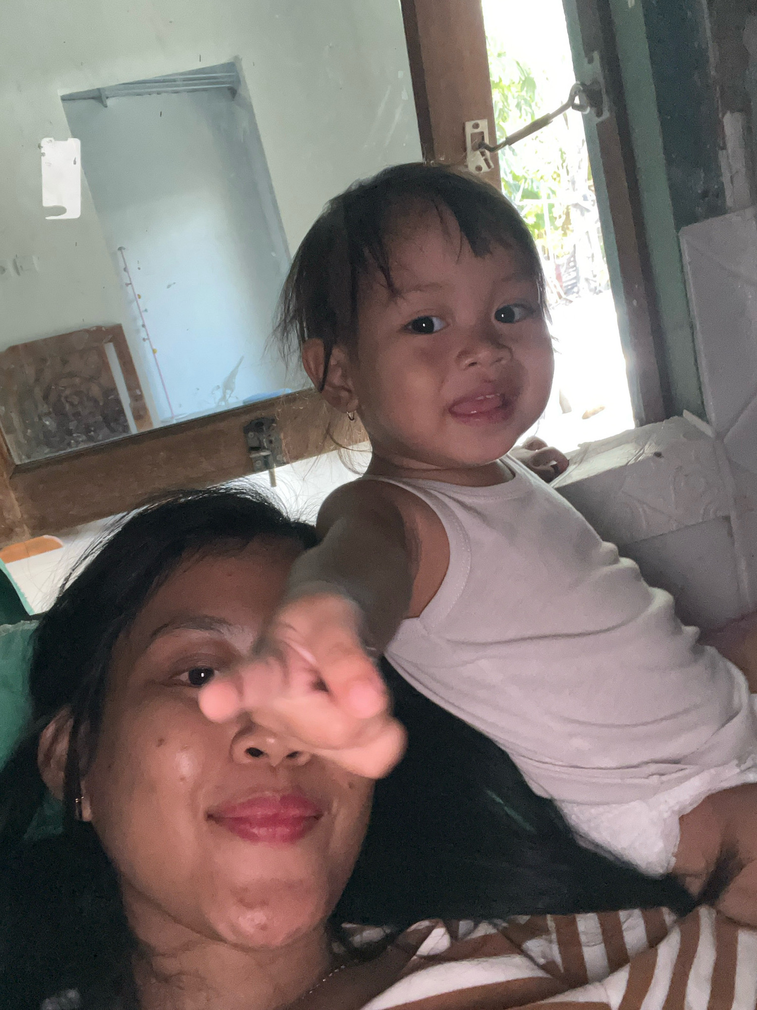An indoor, close-up, selfie-style image shows a woman and a toddler. The woman, in the foreground, has dark hair and is looking at the camera with her right hand extended forward, obscuring part of her face. Her lips are painted pink. The toddler is in the background, wearing a white tank top and a diaper. The toddler is looking at the camera and sticking out their tongue. The background shows a doorway with natural light streaming in, suggesting it is daytime. There is a mirror visible on the left side of the image, reflecting part of the room. The overall environment appears to be a residential setting, likely a home, in Kecamatan Pati, Indonesia.