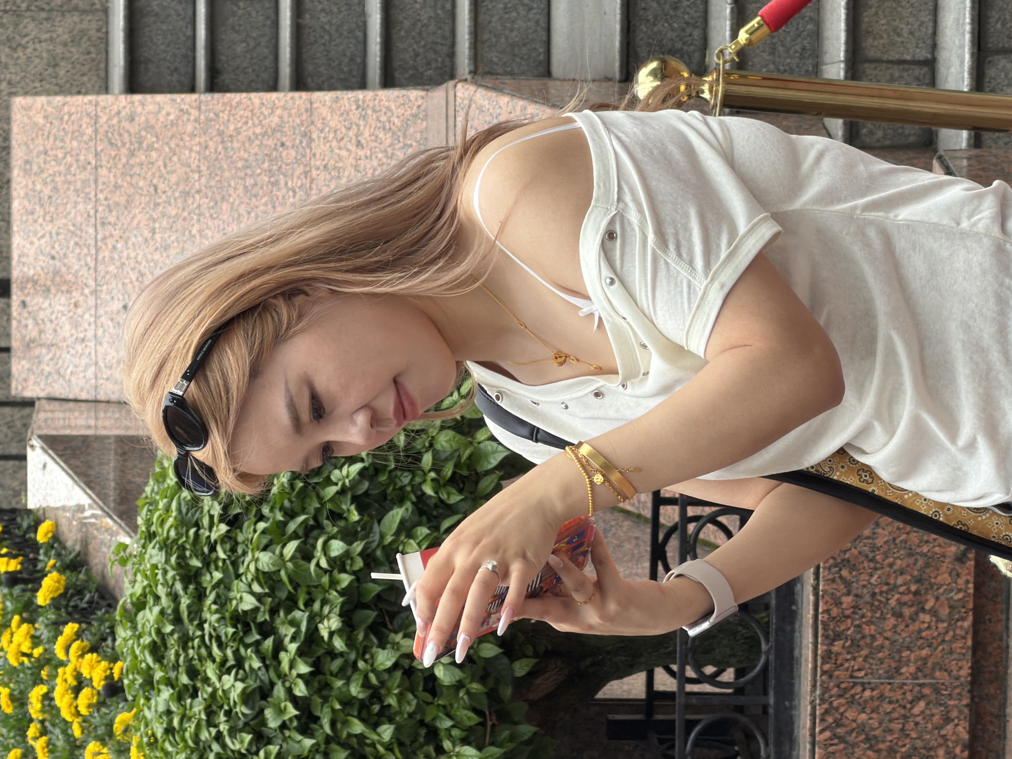 A young woman with blonde hair and sunglasses on her head is standing outside. She is wearing a white off-the-shoulder top, a gold necklace, a gold bracelet, and a ring. She is holding a red and white cup in her hands and looking down at it. In the background, there are green bushes with yellow flowers, a granite wall, and stairs. The scene appears to be in a city, possibly Yuzhong District, China.