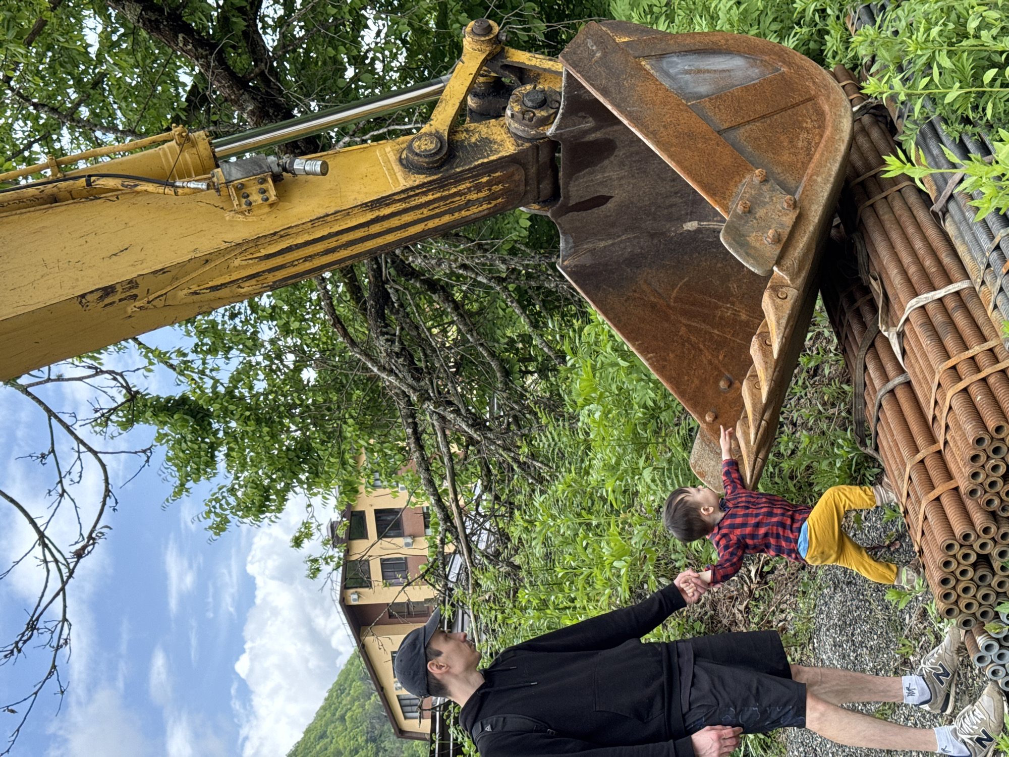 A man in a black hoodie, black shorts, and a black baseball cap is holding the hand of a toddler wearing a red and blue plaid shirt and yellow pants. The toddler is pointing toward the bucket of a yellow excavator. A bundle of rebar lies next to the excavator. A building with brown walls and black windows can be seen in the background, along with a hillside covered in trees. The sky is blue with scattered clouds. The location appears to be Krasnaya Polyana, Russia.