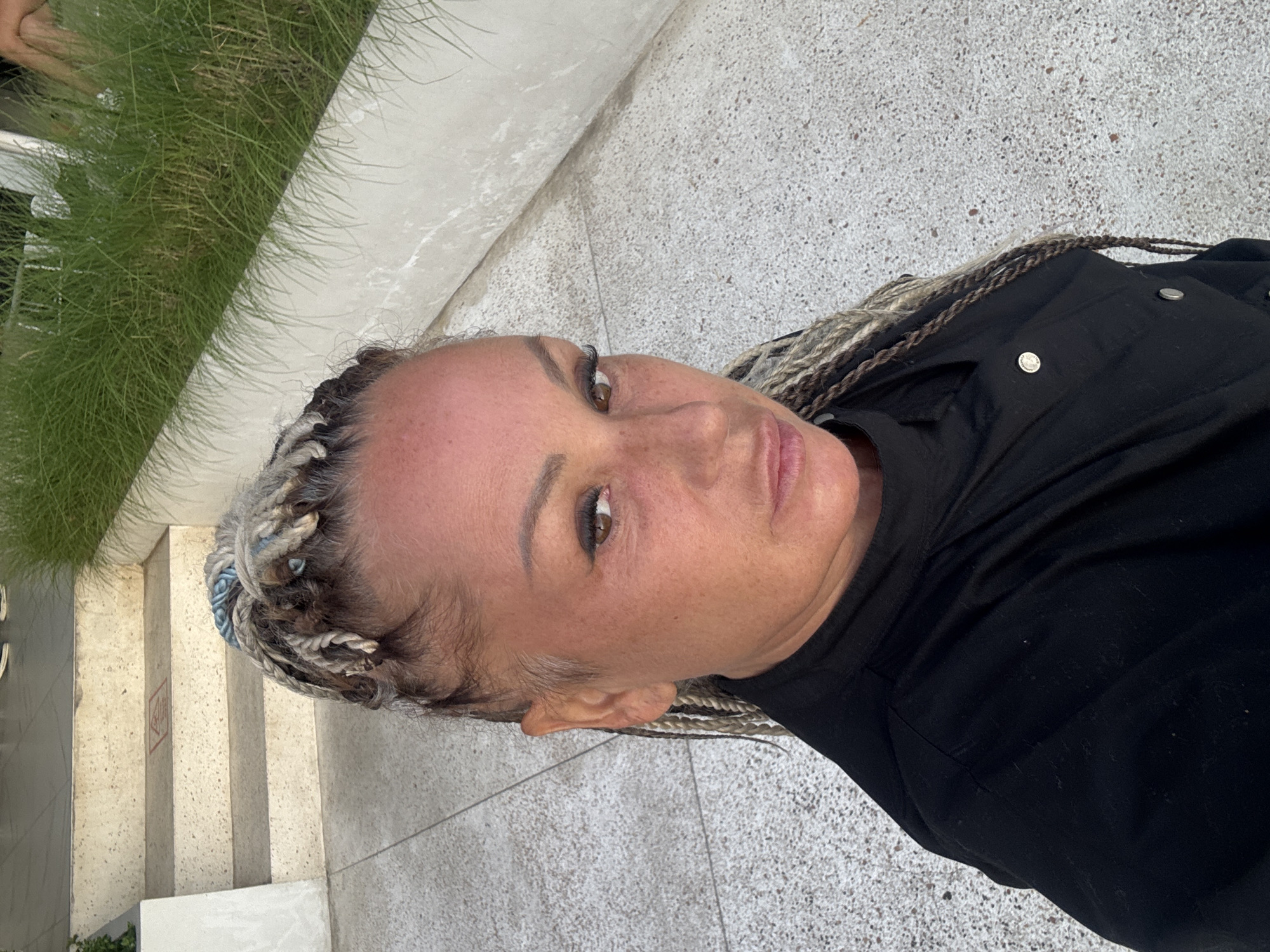 A woman stands outside, possibly in Kedisan, Indonesia. She wears her blonde braided hair up, along with a black shirt. She is near a set of outdoor stairs, and a concrete walkway. Green vegetation is visible next to the stairs.