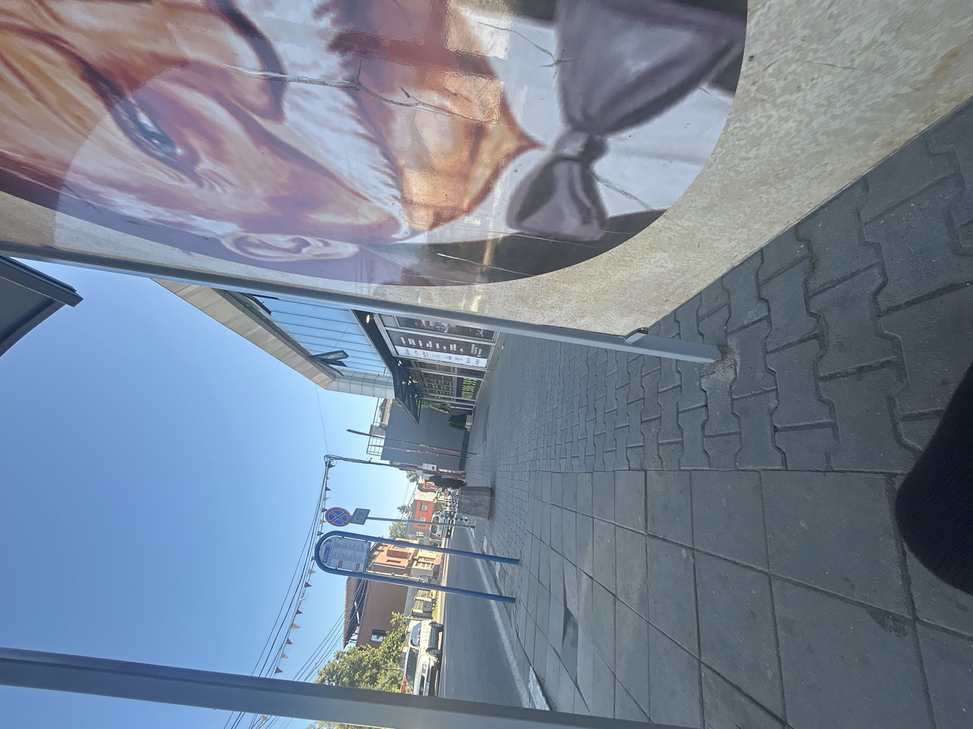 A low-angle, wide shot captures a street scene with a sidewalk in the foreground and a building on the right. In the extreme foreground, a large portion of a weathered, circular advertisement featuring a close-up of an older man's face with a mustache and bowtie is on the right. The man is wearing a dark suit and a white shirt, with a prominent bowtie. The advertisement is positioned at an angle, with its bottom edge extending out of frame.
The sidewalk is paved with interlocking gray pavers and has some slight imperfections. To the left of the sidewalk, there is a street with a white line marking the edge. Several cars are visible on the street, including a silver sedan parked on the left side. A bus stop sign with a blue shelter and a timetable is also present on the left side of the sidewalk, along with a red and white circular road sign indicating a prohibition.
Further down the street, a modern building with a glass facade and several windows is visible. The building has a sign with text that is partially obscured by the advertisement. A few trees are visible behind the buildings on the left side of the image. The sky is clear and blue, indicating a sunny day. The overall scene suggests an urban environment, likely a commercial or residential area.