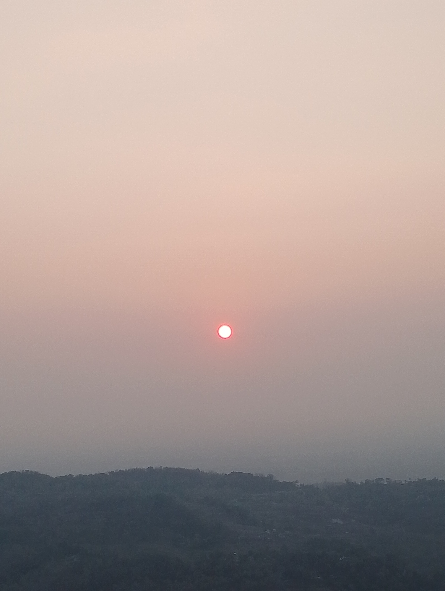 The image depicts a hazy sunset over a landscape of rolling hills, likely in or near Srimulyo, Indonesia. The sun, a small, bright red orb, is positioned in the upper-middle portion of the frame, partially obscured by atmospheric haze. The sky transitions from a pale pinkish hue at the top to a more muted gray towards the horizon. The foreground is dominated by the silhouettes of densely vegetated hills, their shapes softened by the distance and the atmospheric conditions. The overall impression is one of tranquility and subdued natural beauty.