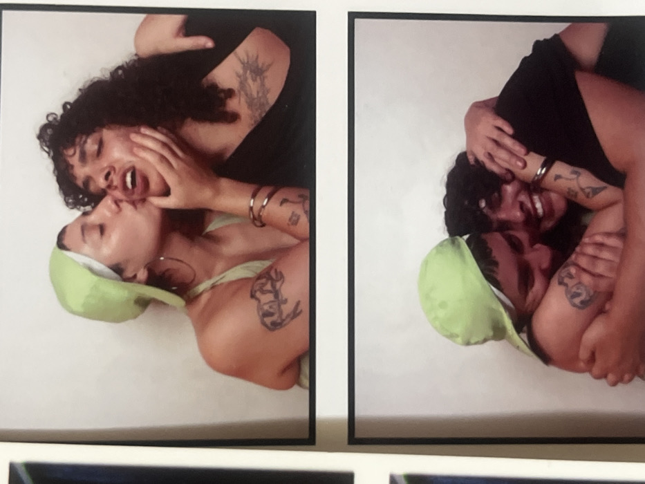 This image displays two vertically stacked photographs, each framed in black on a white border, consistent with photobooth prints, taken within an indoor environment in the city of Buenos Aires, Argentina.
The top photograph features two individuals positioned against a plain white background. The individual on the left, wearing a light green backward cap, a light green halter top, large hoop earrings, and bangles, is depicted kissing the individual on the right near the mouth/cheek area. Tattoos are visible on the left individual's upper right arm, resembling two figures, and another on the inner left forearm. The individual on the right, with dark curly hair and a dark top, has their right hand positioned on the left individual's jawline. Tattoos are visible on the right individual's upper left arm, resembling a tree or plant structure, and another on the inner left forearm.
The bottom photograph shows the same two individuals in a close embrace against a plain white background. The left individual, still wearing the light green cap, has their right arm around the other's shoulders and their left arm wrapped around their back, with their face pressed against the other's shoulder. The right individual is smiling, with their arms wrapped around the left individual's back, and their eyes are open.