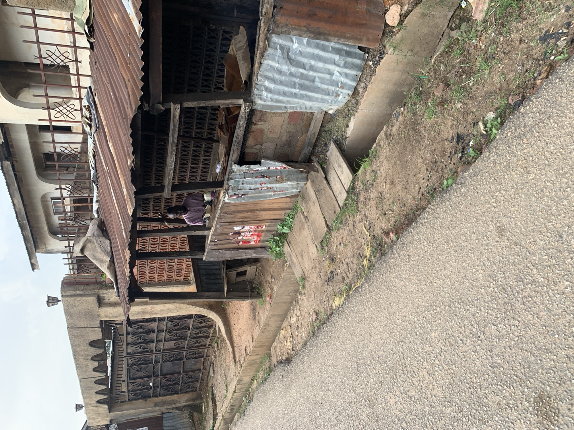 The image shows a street scene in Jos, Nigeria. The view is at an angle, looking down a narrow, paved road. To the left of the road is a row of structures that appear to be market stalls or small shops. These structures are built with a mix of materials, including corrugated metal roofing, wooden beams, and latticed brickwork. Some of the stalls have open fronts revealing goods or storage within.
In the foreground, a concrete drainage ditch runs alongside the road and the stalls. The ditch is partially filled with dirt and overgrown with sparse grass and weeds. The ground beside the ditch is also unkempt, with scattered debris and some rocks.
The buildings to the left of the stalls show more permanent architecture, with arched doorways and decorative metalwork on the windows. Some of these appear to be part of a larger building or complex. A few old-fashioned street lamps are visible on the concrete structures.
The overall impression is one of a bustling, possibly older, commercial area. The time of day appears to be daytime, with the lighting suggesting it is not midday, possibly morning or late afternoon. The weather appears to be fair, with no obvious signs of rain or strong sun. There is no visible text in the image. The scene conveys a sense of everyday life and commerce in an urban environment.