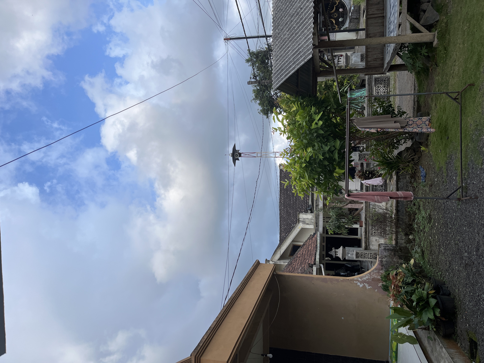 The image depicts a residential area in Jambearum, Indonesia, on an overcast day. A multi-story house with a brown facade is on the left, featuring a wide overhang. To the right, other residential structures with terracotta tiled roofs are visible. In the center of the scene, a tall, slender tower with a speaker at the top stands against the sky. Various power lines crisscross the frame, supported by poles. A small, open-sided wooden structure with a corrugated roof is situated on the right side. In front of the structures, a clothesline holds several items of clothing, including a pink towel and a patterned dress. Lush green foliage and potted plants are scattered throughout the yard, which is covered in gravel and patches of grass. The sky is a mix of blue and white clouds. No people are visible in the image.