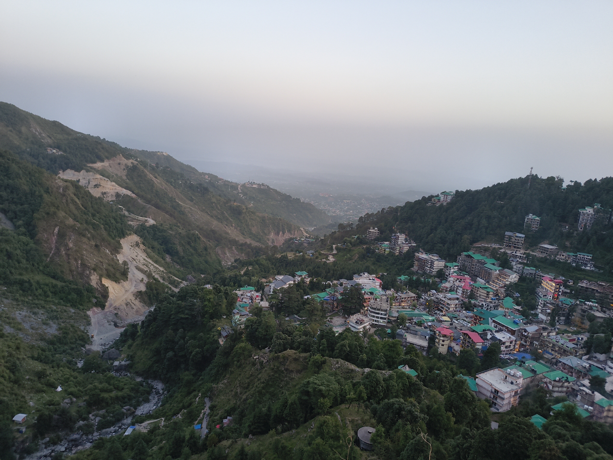 The image depicts an elevated view of a town nestled within a mountainous region of Bhagsu, India. Buildings with green, red, and gray roofs are scattered amongst dense, green foliage on the hillside. In the background, the view extends to the valleys and distant mountains, partially obscured by a layer of haze or fog. A road with signs of excavation or road repair is visible on the left side. The sky is a muted light blue, suggesting dawn or dusk. No people are distinctly visible.