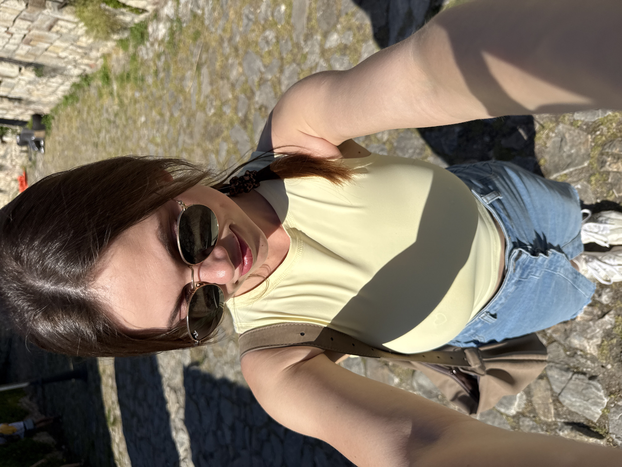 A woman is taking a selfie outdoors. She is wearing sunglasses, a light yellow top, and blue jeans. She has a brown bag slung across her body. The background shows a stone pathway and wall, with some grass and greenery visible. Based on the environment, this photo was taken in Belgrade, Serbia.