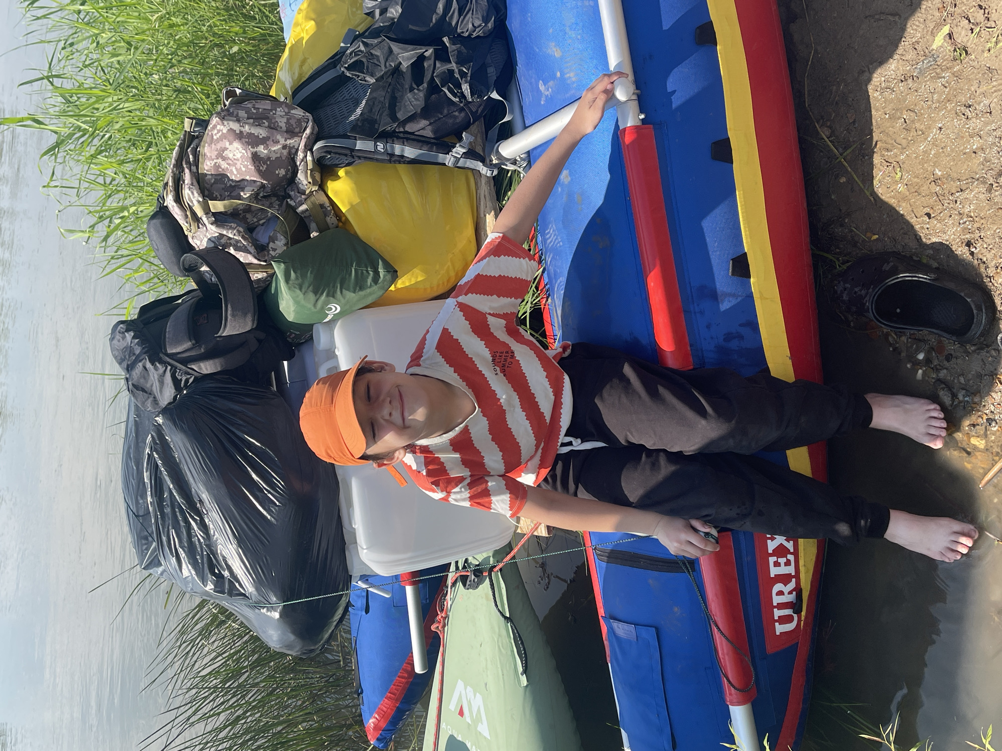 A young person wearing an orange baseball cap and a striped t-shirt sits on a blue and red inflatable raft, facing the camera with a smile. Their legs are bare and dangling in the water. The raft is loaded with various bags, including a large black trash bag, backpacks, and a yellow waterproof bag. The raft is positioned at the edge of a body of water, with tall green grass growing along the bank. The ground is muddy. In the background, the water surface is visible under a clear sky. The scene appears to be a natural outdoor setting, possibly a riverbank or lake shore. The location is identified as Nezhinka, Russia.