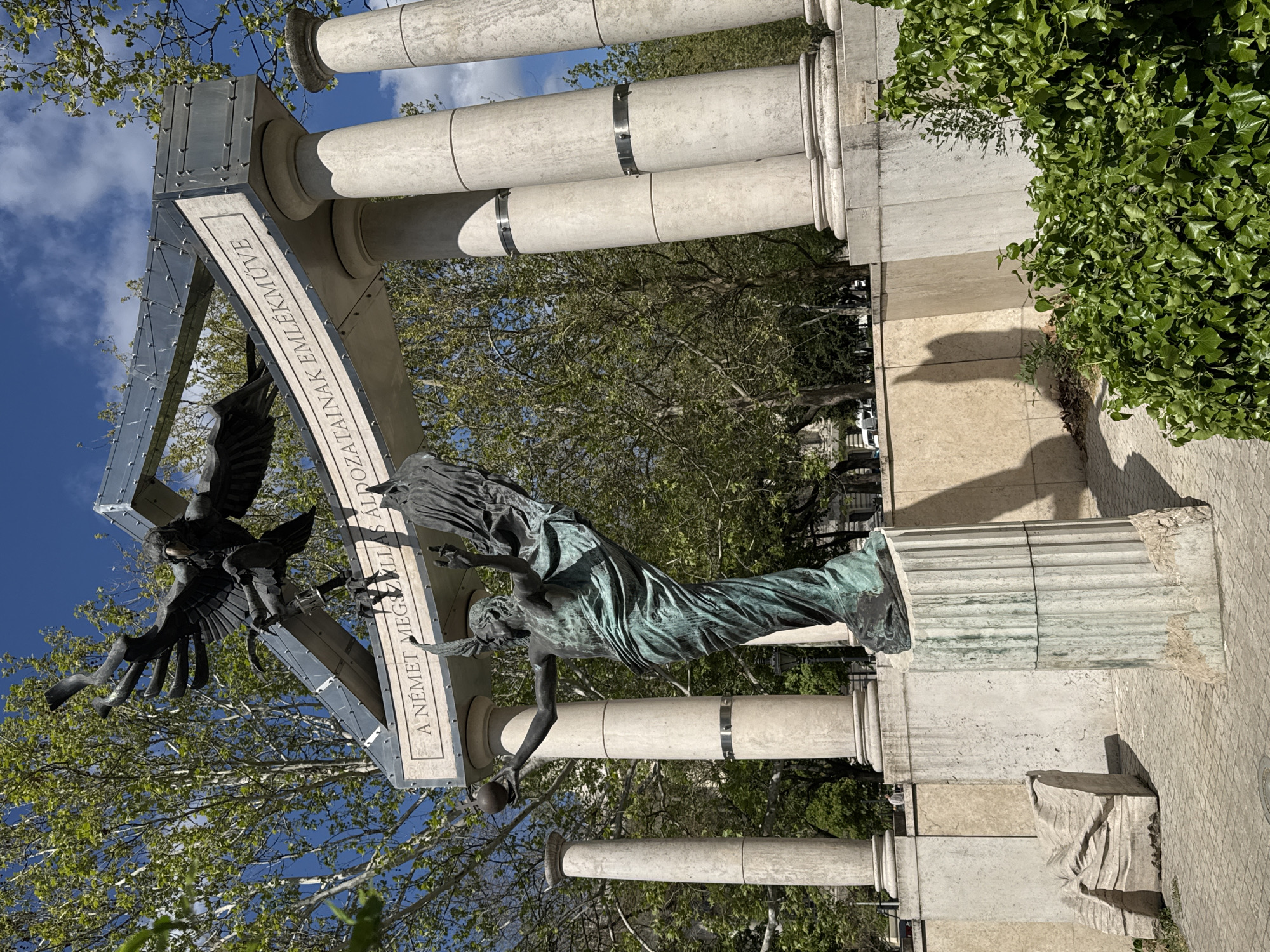 The image displays a monument in Budapest, Hungary, under a bright, clear blue sky with sparse white clouds, suggesting mid-day. The monument features a central bronze statue of an angel-like figure, patinated green, depicted in flowing robes. Its left hand holds a spherical object, while its right hand is outstretched in a gesture of distress or resistance. Above and slightly to the left, a dark, eagle-like bird sculpture with outstretched wings and claws swoops aggressively towards the figure.
These statues are framed by a prominent, modern arch-like structure made of metallic panels with sharp angles. This structure is supported by several classical, light-colored stone columns, some of which feature metallic bands. Inscribed on the arch in white lettering against a dark background is the Hungarian text: "A NÉMET MEGSZÁLLÁS ÁLDOZATAINAK EMLÉKMŰVE," which translates to "Monument to the Victims of the German Occupation."
The background shows lush green trees with fresh foliage, indicative of spring or early summer. The ground is paved with light-colored stones, and a dense growth of green ivy covers part of a wall or base structure to the right. The strong sunlight casts distinct shadows, highlighting the architectural and sculptural details. The scene portrays a powerful symbolic struggle, commemorating a historical tragedy.