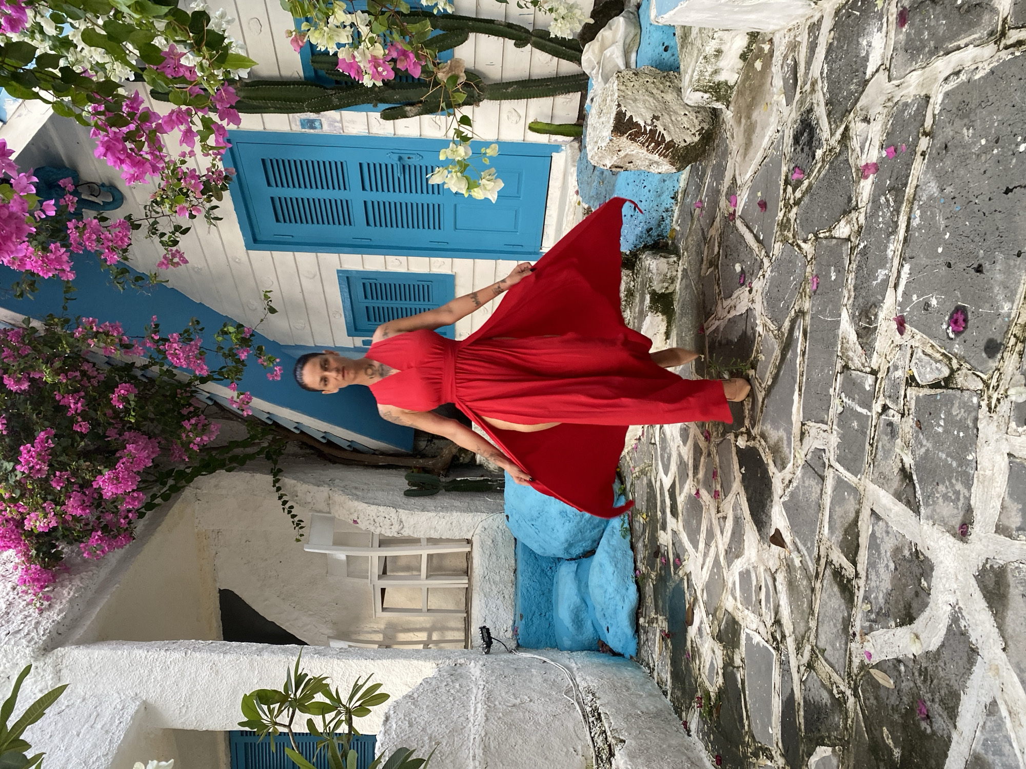 A woman in a red dress stands on a stone pathway, holding the dress out. Behind her is a white building with blue doors and accents, adorned with bougainvillea. The scene appears to be an exterior location in Phu Thuy Ward, Vietnam.