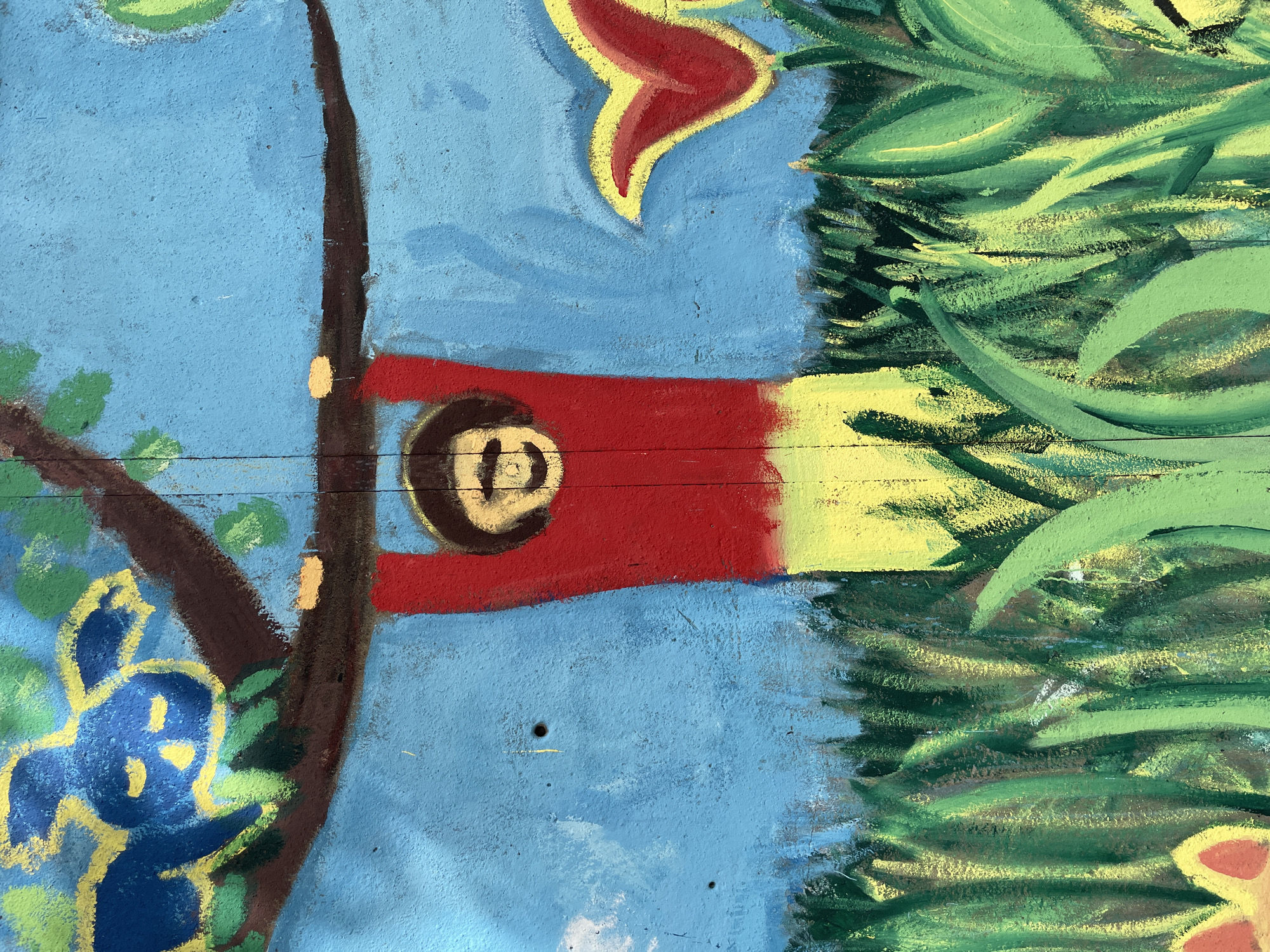 This is a close-up, eye-level photograph of a colorful mural painted on a textured, light blue wall. The mural depicts a simplified, stylized scene with elements of nature and a human figure.
Dominating the center is a brown tree branch extending horizontally across the frame, from which a figure is hanging. The figure is depicted with a round head featuring a simple smiley face drawn in brown and yellow. The body is a rectangular shape painted in bright red from the neck down to the waist, transitioning to a bright yellow from the waist to the bottom edge of the frame. The figure's hands are gripping the brown branch.
Above the branch, on the left side of the image, is a blue and yellow creature resembling a stylized animal, possibly a bird or a monkey, with large, closed eyes and floppy ears. It is resting on the branch amidst green foliage.
Below the figure and the branch, the lower half of the mural is filled with green and yellow brushstrokes representing tall grass and vegetation. Two flowers are visible: one on the right, a red and yellow tulip-like flower, and another on the far left, a yellow and orange flower peeking out from the grass. The sky is a solid, bright blue, with some faint white brushstrokes suggesting clouds.
The wall surface appears rough, with visible texture and some minor imperfections, including two small holes. The artwork has a childlike, whimsical quality, likely created with chalk or pastel-like paints due to the powdery texture and blending of colors.
The mural is located on a wall in Bremerhaven, Germany, specifically at the Kinderspielplatz (children's playground) at Lange Straße 41, adjacent to the kindergarten.