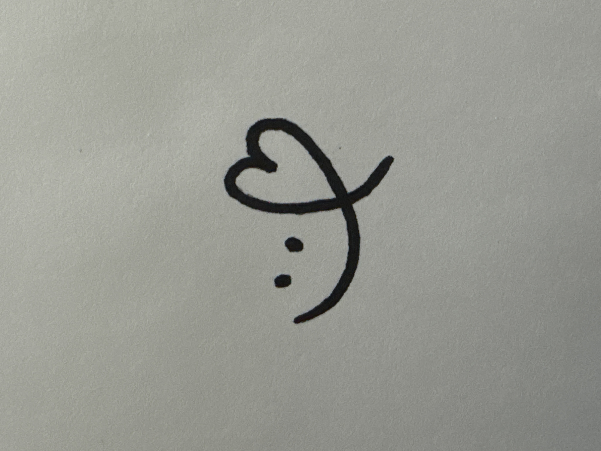 A minimalist black drawing on a grey textured surface depicts a smiley face with a heart to the right. The smiley face consists of a curved line for the mouth and two dots for the eyes. The heart is drawn with a single continuous line that also forms part of the smiley face's mouth. The drawing is centered on the surface.