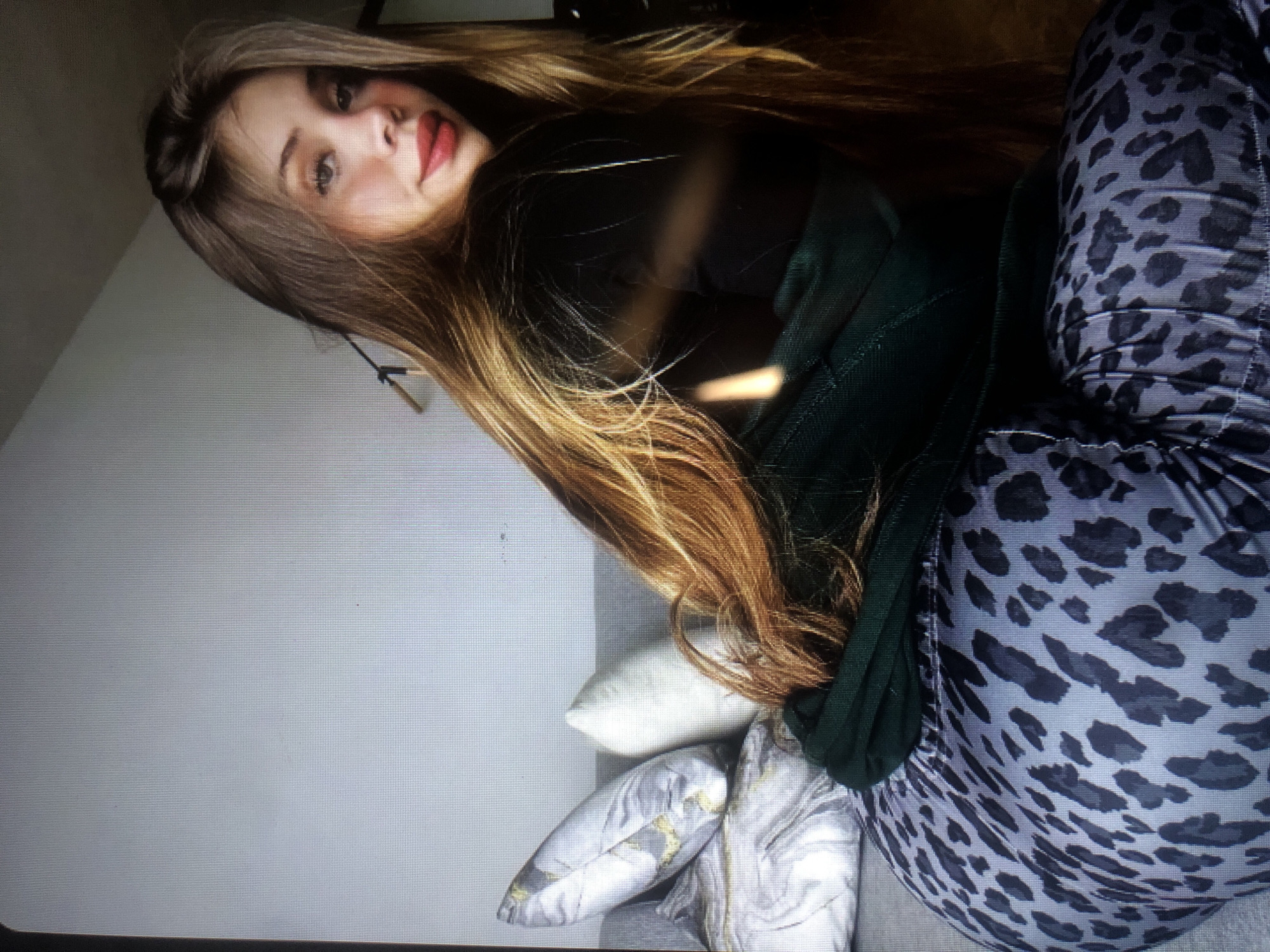 The media file is a portrait of a young woman lying down, with her head propped on pillows. She has long, blonde hair that falls around her shoulders. She is wearing a dark green long-sleeved top and patterned leggings with a leopard print. Her makeup includes bold red lipstick and dark eyeliner. The setting appears to be indoors, possibly a living room, with a light-colored wall in the background and several decorative pillows. The lighting suggests it could be daytime, as there's a window or light source casting a soft glow. The overall mood is relaxed and casual. There is no visible text in the image.
