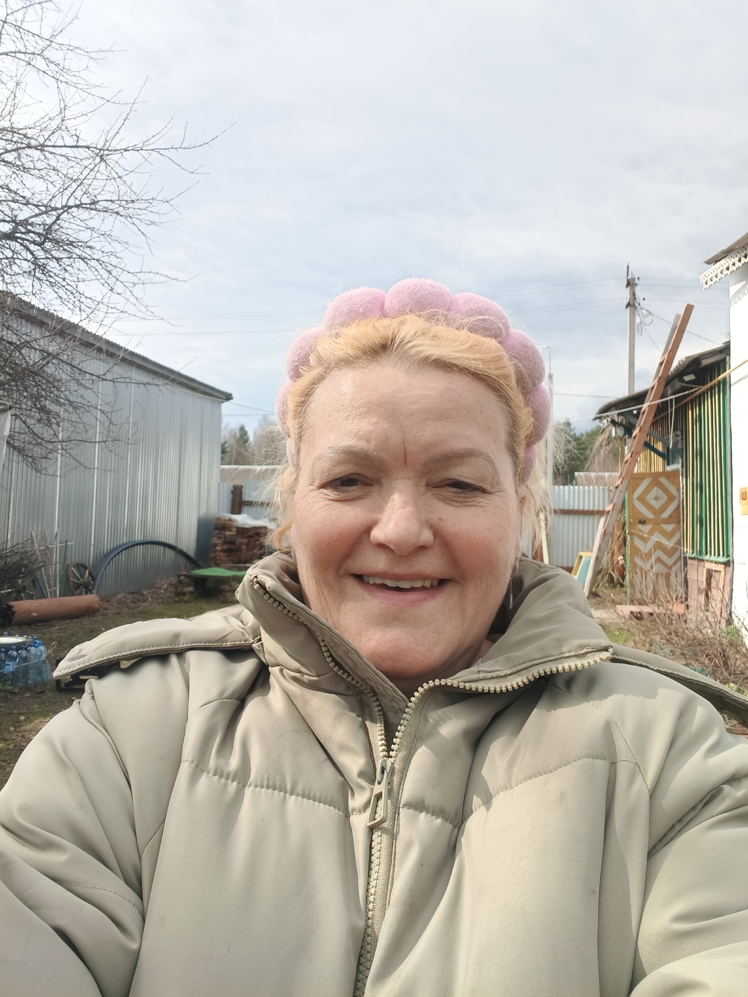 A smiling woman, appearing middle-aged, is captured in a close-up selfie outdoors in Красный Маяк, Russia. She has blonde hair and wears a distinctive pink, fluffy headband, along with a warm, light olive/beige padded winter jacket. Her expression is one of happiness, characterized by a broad smile.
The setting is a rural yard, likely in late winter or early spring, given the bare branches of a tree to the left of the frame. The sky is overcast with light grey clouds. Behind the woman, to her left, stands a corrugated metal shed or outbuilding. Below it, various items are scattered, including an old wooden wagon wheel, a large curved metal sheet, a terracotta pipe, and a cluster of plastic bottles. To her right, another structure is visible, possibly wooden, with vertical green slats and a small door decorated with a yellow and white diagonal pattern. A wooden ladder leans against this building. A utility pole with power lines extends upwards into the sky. Fencing is also visible in the background. No discernible text is present in the image.