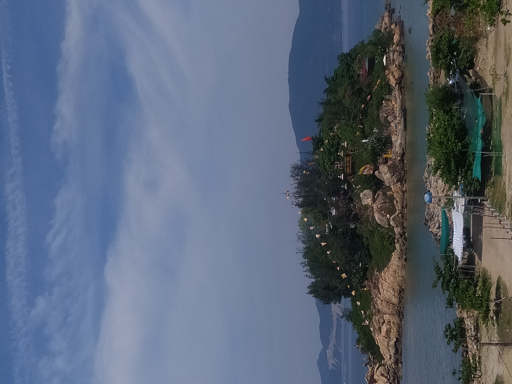 A high-angle, full-shot photograph shows a scenic view of a small, rocky island covered in lush green vegetation and trees. The island is situated in the middle of calm, blue water, with a larger landmass and mountains visible in the distance under a bright, partly cloudy sky. The island features several structures, including what appears to be a small Buddhist shrine or temple complex with statues and prayer flags. A red flag flutters from a pole on the island. Along the shoreline of the island, there are exposed rocky formations. In the foreground, the shoreline is lined with green trees and vegetation, and there are some simple structures with tarps or awnings set up, possibly for shade or a small market. A dirt path leads towards the structures. The scene depicts a serene coastal landscape.