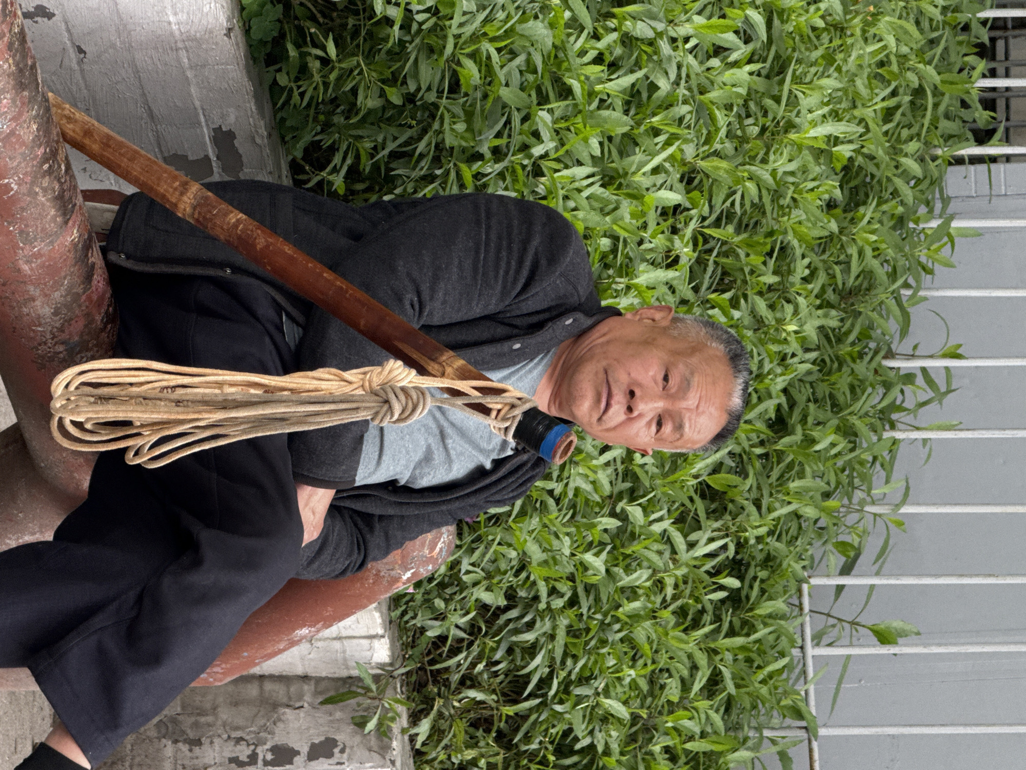 An adult male of East Asian descent, with short grey hair, is seated outdoors in Jiulongpo District, China. He wears a dark grey zip-up jacket over a light grey t-shirt and dark trousers. He faces forward, looking directly at the camera with a neutral expression. A long, dark brown wooden staff is angled diagonally in front of him, resting against his left side. The upper end of the staff is wrapped with blue and black material. A coil of beige rope is tied around the staff near the top. The individual is seated on a reddish-brown painted concrete or metal structure. Behind him is dense green foliage, backed by a light grey wall with white vertical bars. A lower concrete wall with peeling white paint is visible below the foliage.