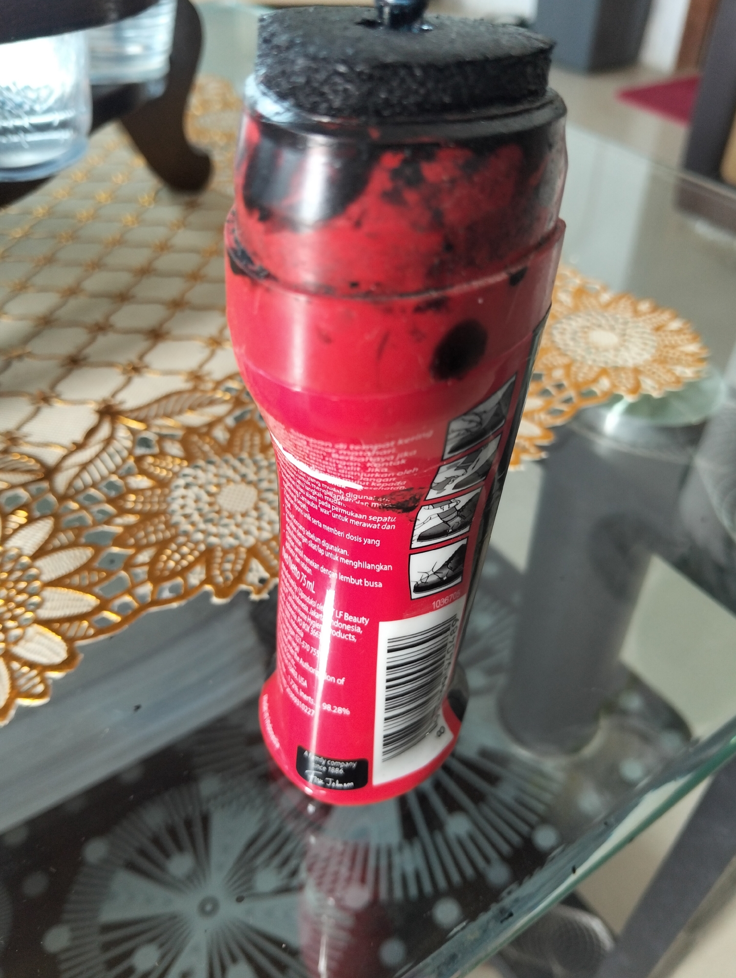 A red shoe polish bottle with a black applicator cap is on a glass table. The bottle has some black smudges on its surface and a label with text and illustrations of how to use the product. The label also shows a barcode. In the background, a decorative placemat with a gold and white pattern is visible. The environment appears to be indoors, possibly a home or office, with other objects and furniture blurred in the background. The city of Julicotmeurak, Indonesia, is mentioned on the label, indicating the location of manufacture or distribution.