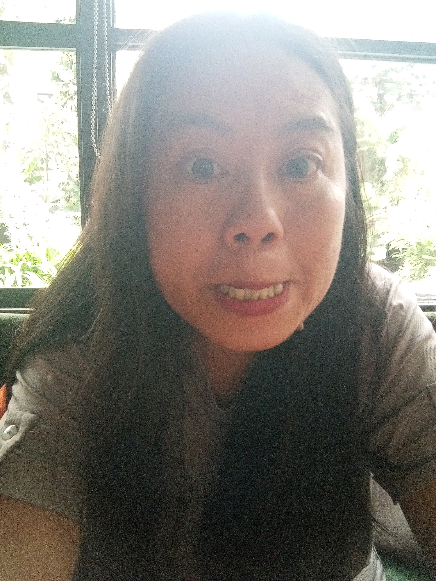 A medium shot shows a woman with long, dark hair and fair skin. She is wearing a grey t-shirt with a button and a folded cuff on the left sleeve. Her eyes are wide open, and her mouth is slightly open, revealing her teeth. The woman is looking directly at the camera. The background is out of focus, showing a window with what appears to be greenery outside. The lighting is bright, with a strong glare coming from the top of the image. The image appears to be a selfie. The scene is likely an indoor location with natural light.