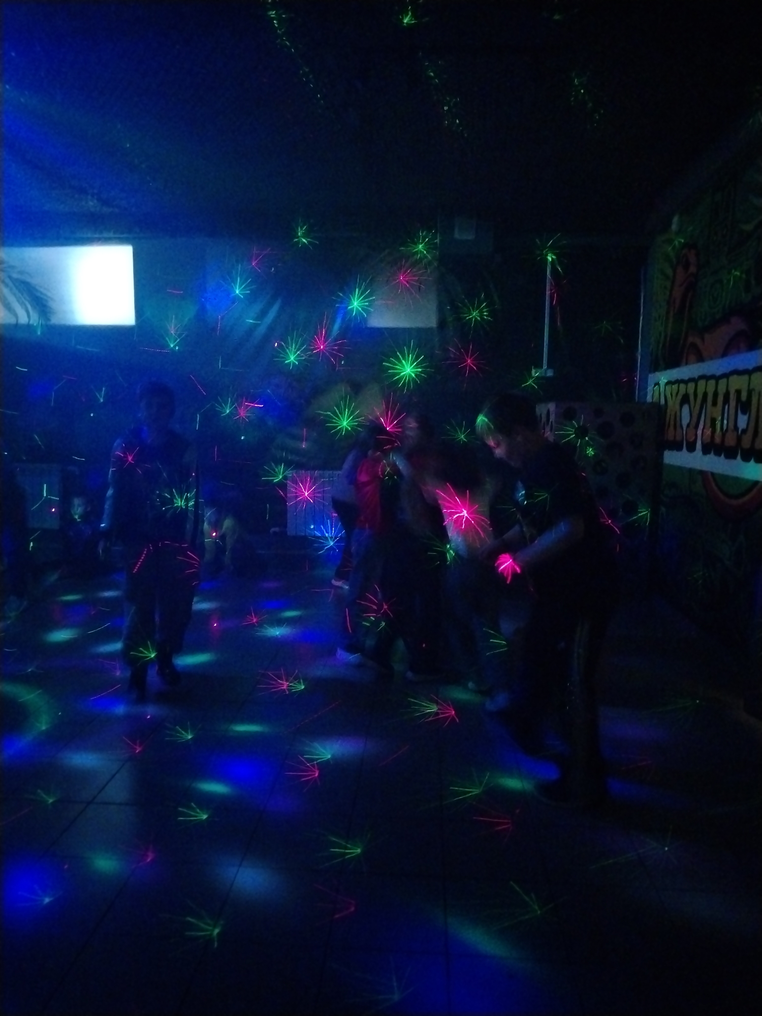 The image shows a dark interior space, likely a recreational or entertainment venue, in Rusanovo, Russia. Multiple people are present, standing and interacting amidst a dense display of laser lights. The laser lights project patterns of green, blue, and pink stars onto the floor and walls. A screen or light fixture emits a bright, rectangular glow in the background. The floor appears to be tiled. Some signage with lettering is visible on a wall. The overall lighting is dim, creating a nightclub-like atmosphere.