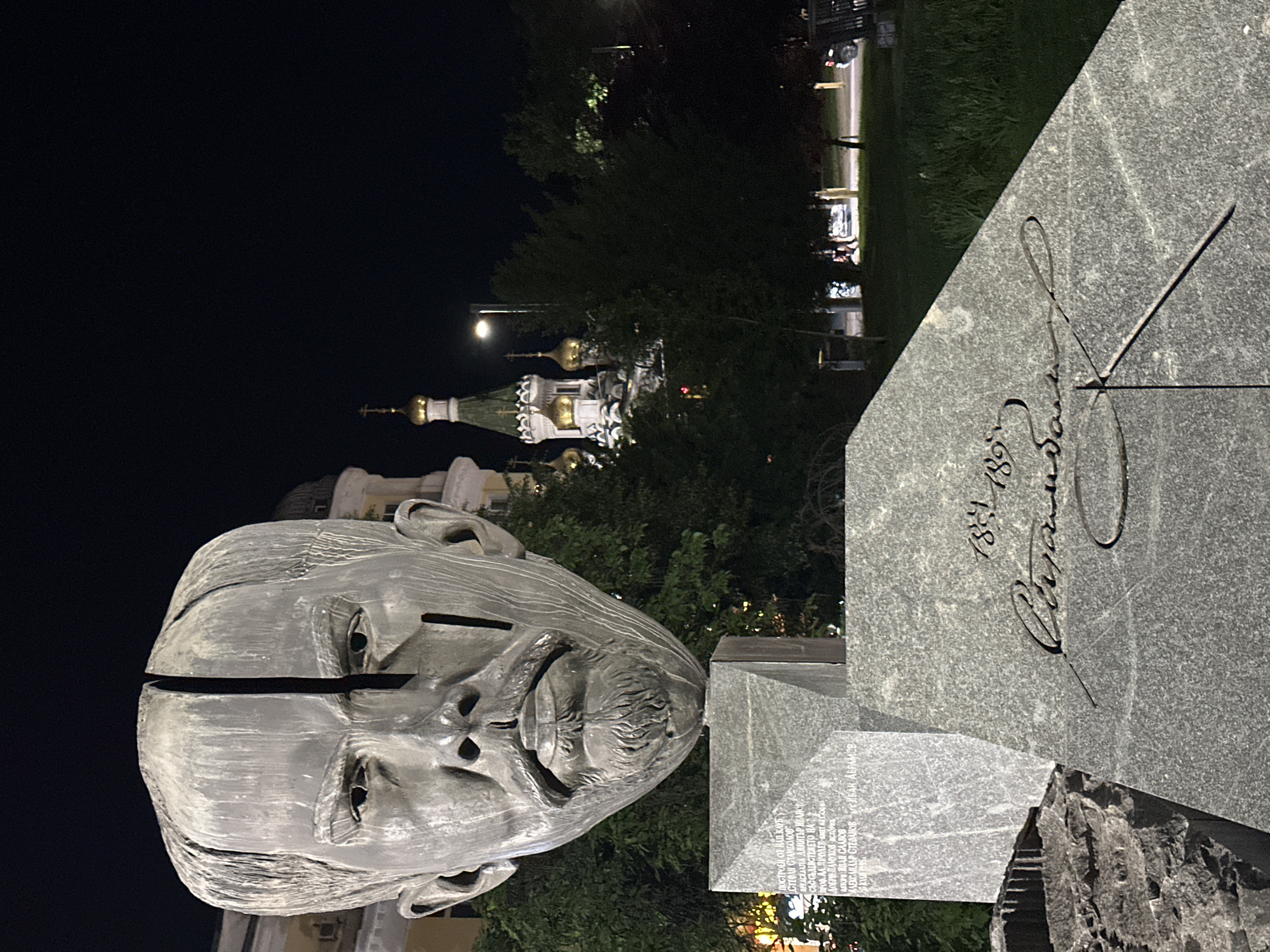 The image displays a monument at night in Sofia, Bulgaria, featuring a large, artistic sculpture of a human head and a granite base, with a church visible in the background.
The main subject is a large, metallic sculpture of a human head, possibly male, with a distinctive vertical split running down the middle of the face. The head rests on a textured, rough-hewn stone pedestal, forming part of a larger granite monument.
Adjacent to the head sculpture is a large, polished grey granite slab, angled upwards. Etched into this slab is a stylized signature or name, along with the dates "1851 189)". Below this, on a lighter grey granite section of the monument's base, is an inscription in Bulgarian Cyrillic. Visible phrases include "ПОСТРОЕН ОТ НАД КЛУБ" (Built by above Club), "СТЕФАН СТАМБОЛОВО" (Stefan Stambolov), "ПРЕДСЕДАТЕЛ ДИМИТЪР ИВАНОВ" (Chairman Dimitar Ivanov), "СЪС СЪДЕЙСТВИЕТО НА" (with the assistance of), and other names like "ПИЧУЛЕВ-КМЕТ НА СОФИЯ" (Pichulev - Mayor of Sofia) and "АЛЕКСАНДЪР СТЕФАНОВ". This suggests the monument commemorates Stefan Stambolov and others involved in its creation or related historical events.
In the background, partially obscured by trees, an illuminated Orthodox church with white walls, multiple green-tiled roofs, and golden onion-shaped domes topped with crosses is visible against the dark night sky. The scene is lit by artificial lights, including streetlights, indicating it is nighttime. Trees and grassy areas surround the monument, suggesting a park or public square setting. No specific activity is taking place; the image captures a static outdoor scene.