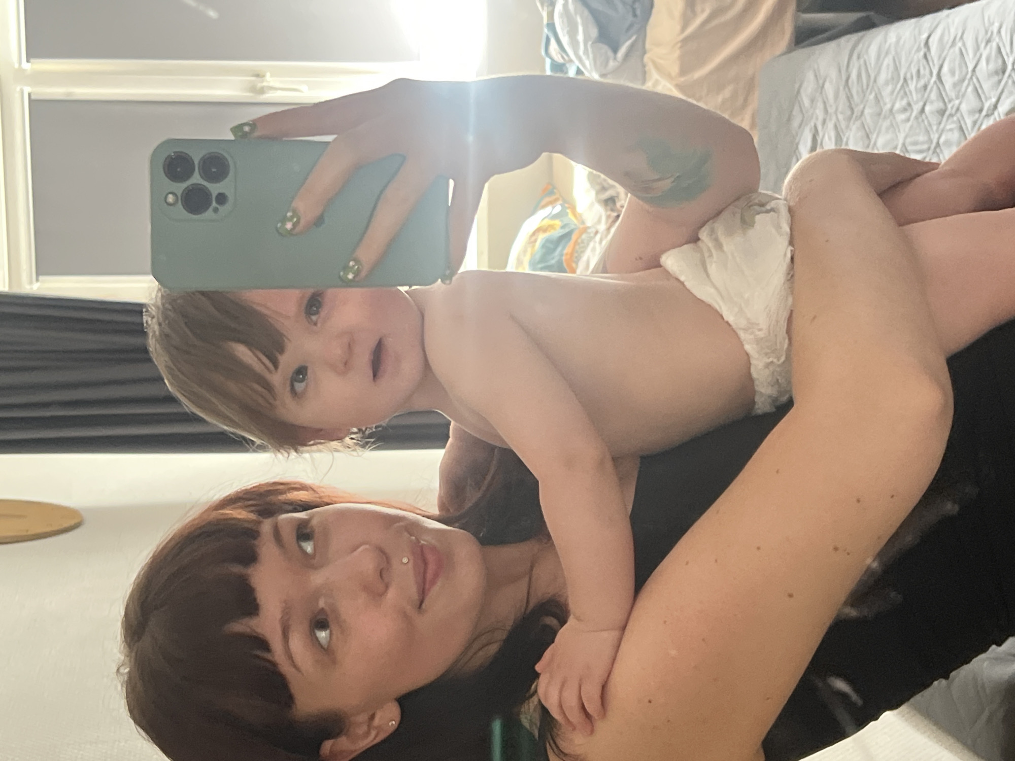 A selfie is taken by a young woman who is lying down, holding a baby. The woman has dark brown hair with bangs and is wearing a black shirt. She has a nose ring and an earring. The baby, who is lying on the woman's chest and torso, is wearing a diaper. The baby has light brown hair and is looking at the phone. The woman is holding a light blue phone in her right hand, with her nails painted green with floral designs. The background appears to be a bedroom, with a bed and some decor. Sunlight is streaming in from the left, creating lens flare on the phone and illuminating the subjects. The overall mood is intimate and casual, suggesting a moment of connection between mother and child.