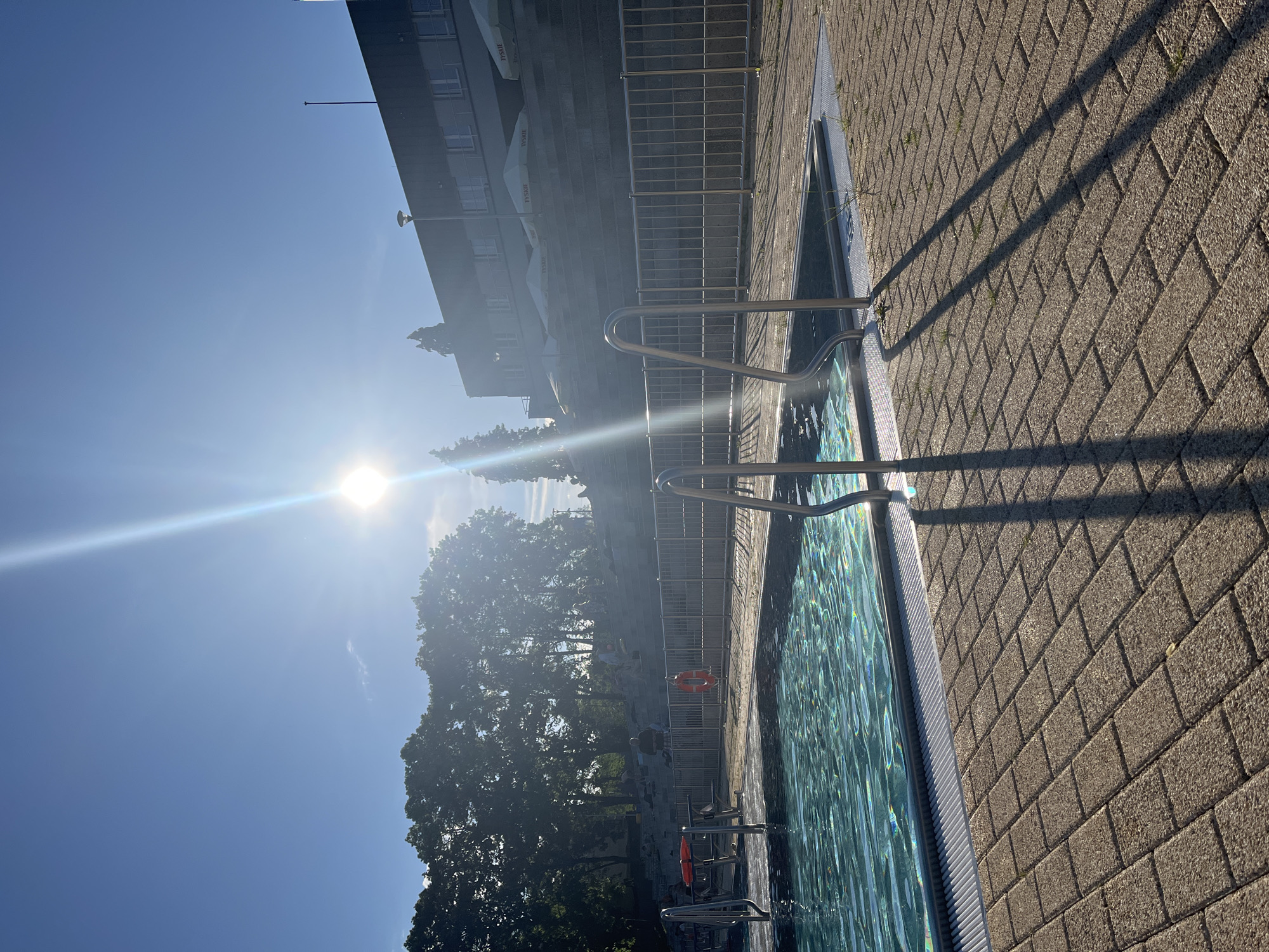 This is an outdoor scene depicting a swimming pool area under a clear blue sky, presumably in the city of Unknown, Unknown. The image captures a bright, sunny day, with the sun prominently visible in the upper right quadrant, casting strong lens flare and long, distinct shadows towards the lower left, indicating either early morning or late afternoon.
In the immediate foreground, light brown/grey interlocking concrete pavers form the ground surrounding the pool. Long, dark shadows of the pool's stainless steel handrails stretch across these pavers, interspersed with small patches of green weeds.
The central feature is a rectangular swimming pool filled with clear, turquoise water, exhibiting ripples on its surface that reflect the sunlight. Two sets of stainless steel handrails are visible for pool entry/exit, one on the left and one on the right.
Behind the pool, a silver-colored metal fence with a grid pattern acts as a barrier, separating the pool deck from a raised terraced area. On the left side of the pool, near the fence, a red life buoy is mounted on a stand, and another orange life buoy hangs on the metal fence.
The background comprises two main elements: on the left, a dense cluster of green deciduous trees with sunlight filtering through their leaves. On the right, a multi-story building with a dark roof and a lighter grey facade featuring multiple windows. Below this building, on a raised platform, several white parasols with "TYSKIE" branding are visible. A streetlamp pole stands near the building. Between the trees and the building, a terraced seating area made of large stone steps or platforms is visible. Several indistinct figures are seated or standing on these steps, appearing to be relaxing or sunbathing.