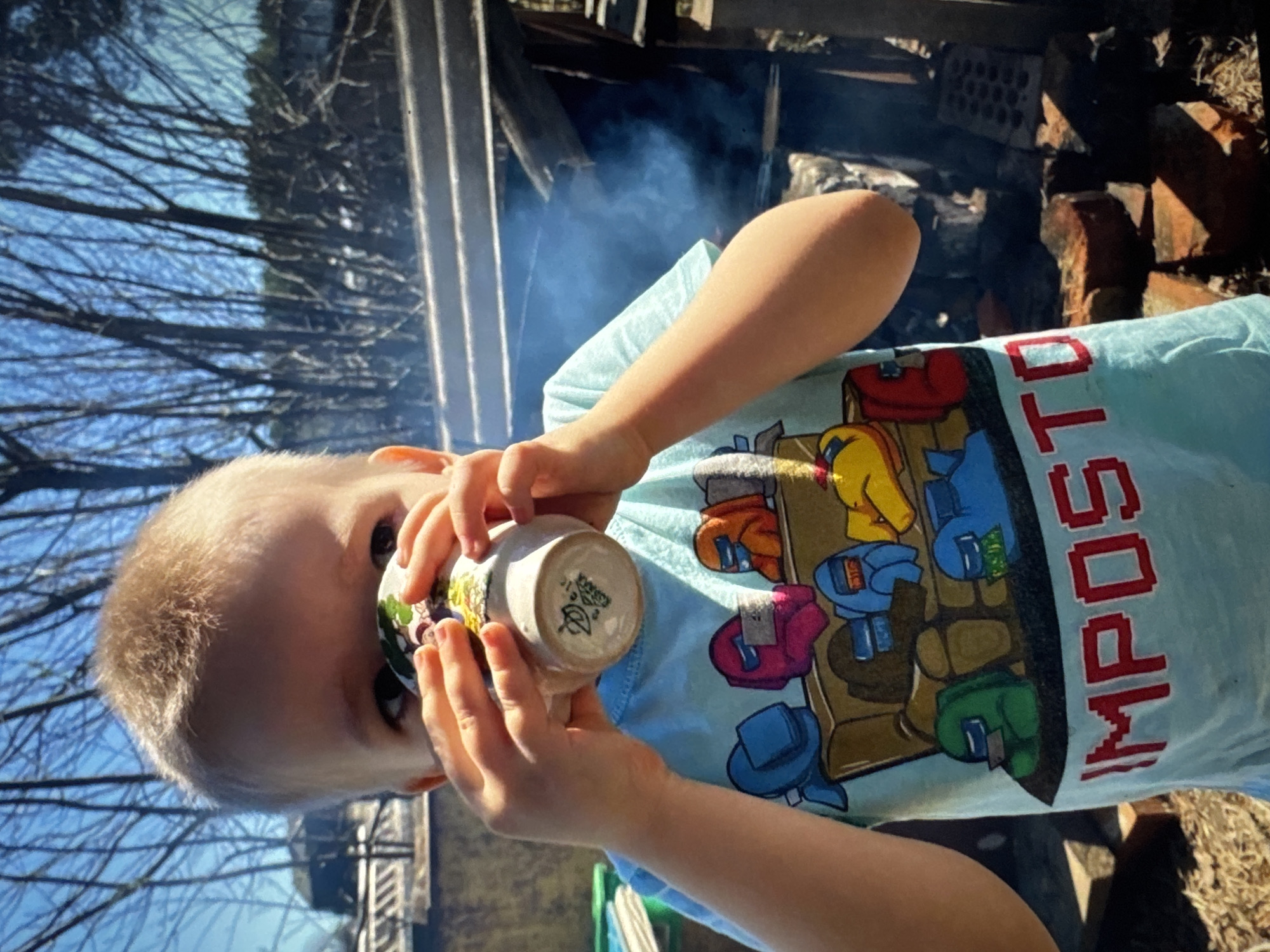 A young child is drinking from a mug, seemingly outdoors. The child has blond hair and is wearing a light blue t-shirt with an "Among Us" themed graphic. The background shows trees without leaves, a fence, and some smoke, suggesting a possible outdoor fire. There are bricks visible in the background. The image does not confirm the specific location in the city of Solikamsk, Russia.