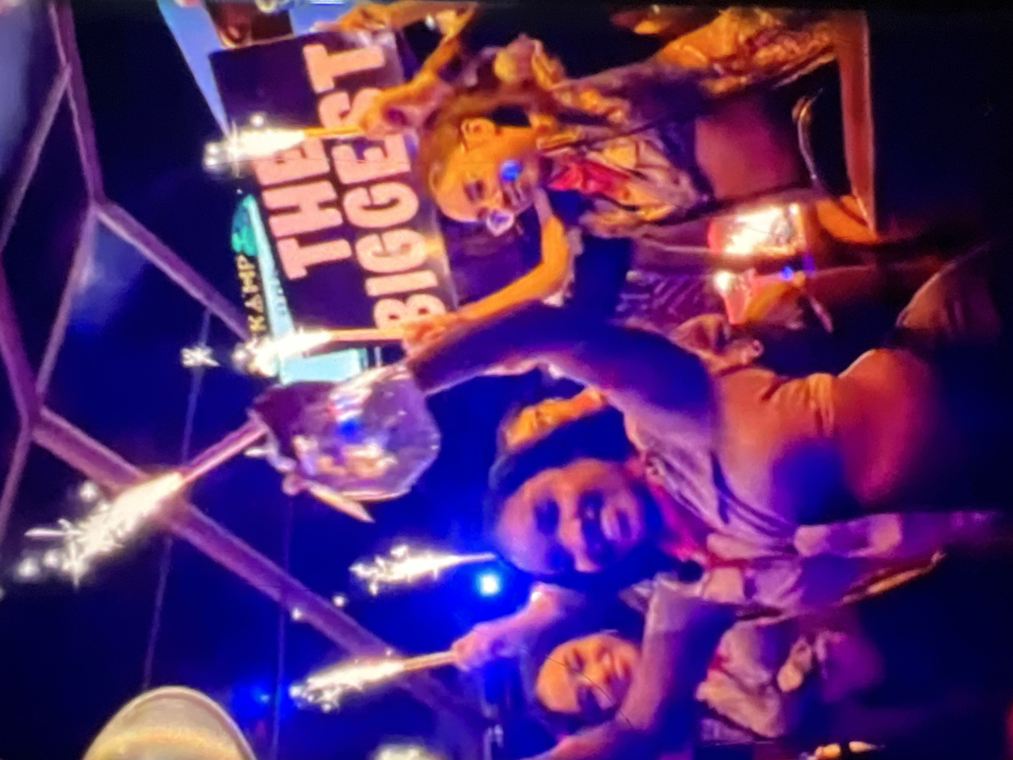 The image shows a group of people celebrating indoors, with sparklers and a sign that reads "THE BIGGEST". The setting appears to be a nightclub or party venue with dim, colorful lighting. The people are dressed casually and appear to be enjoying themselves. One person in the foreground is holding up a sign, while others are holding sparklers. The atmosphere is festive and energetic. The image quality is poor, making it difficult to discern finer details. While there is a neon sign in the background that says "KAMP", this does not specifically indicate the location in Lagos, Nigeria, as "Kamp" could be a generic name for a venue or event.