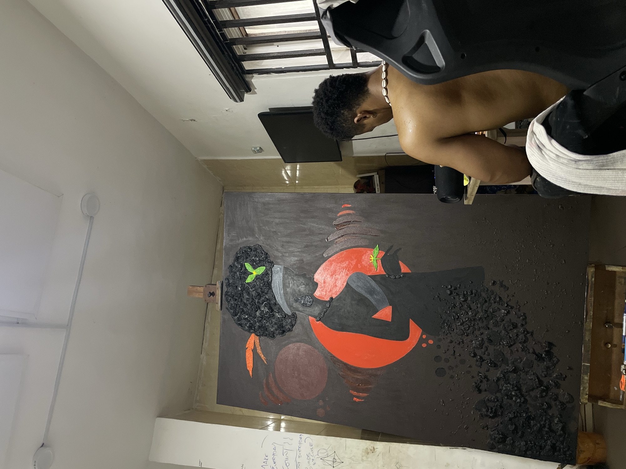 The image shows a person with dark hair and a shell necklace, partially visible and shirtless. They appear to be standing near a large abstract painting depicting two figures in black and red against a dark gray background, with what appears to be textured black material adhered to the canvas. The painting is resting on an easel in a room with white walls. Near the painting, there's a sheet with handwritten notes. A television is mounted on the wall. The image appears to be taken indoors in a residential area, possibly a private residence or art studio in Ado, Nigeria.