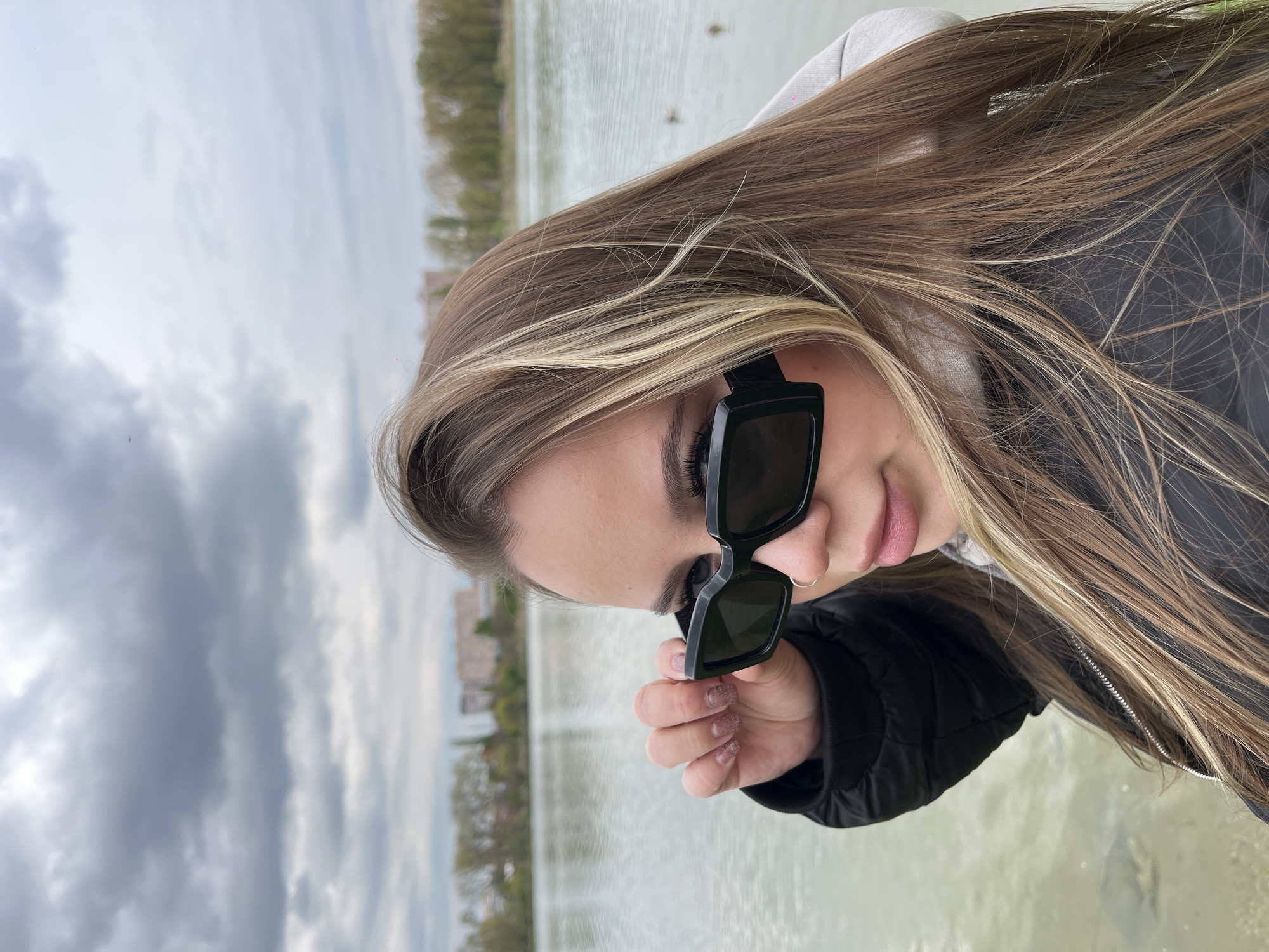 A close-up to medium shot captures a young woman outdoors in Krasnodar, Russia. She is looking downwards and slightly to her right, holding her black, square-framed sunglasses with her right hand just below her eye, as if adjusting them or peeking over them. Her expression is neutral. She has long, straight light brown hair with noticeable blonde highlights, which falls over her shoulders. She is wearing a black puffer jacket over what appears to be a light-colored hoodie. Her nails are manicured with a subtle glittery polish.
The background shows a calm body of water, possibly a lake or large pond, reflecting the overcast sky with a light, greenish hue. In the mid-ground, a subtle fountain spray is visible in the water. Beyond the water, a distant shoreline with trees and residential buildings can be seen under a largely cloudy, grey sky. The overall scene suggests an overcast daytime setting. No text is visible in the image.