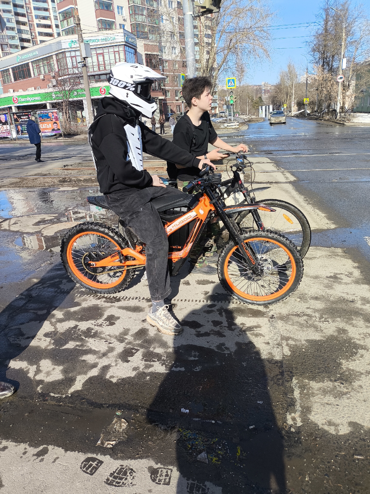 The scene depicts two individuals and their two-wheeled vehicles stopped on a city street in Izhevsk, Russia, specifically near a commercial building complex featuring a "Пятёрочка" (Pyaterochka) supermarket, "БАНК УРАЛСИБ" (Uralsib Bank), and "МЕДСИ" (Medsi clinic).
In the foreground, a male individual is seated on an orange and black "KUGOO" electric motorcycle with knobby tires and orange rims. He wears a white full-face motocross helmet with "100%" branding, a black hoodie over a white chest protector, dark jeans, and light-colored sneakers. His right foot is placed on the damp asphalt. To his right, another male individual stands next to a dark-colored bicycle with an orange front wheel reflector. This individual wears a black t-shirt and dark pants, holding a smartphone in his left hand while resting his right hand on the bicycle's handlebars. Both individuals are partially obscured by long shadows stretching from the left side of the frame.
The ground consists of dirty, wet asphalt pavement with scattered puddles, mud, and visible shoe prints. Tram tracks run across the road surface. In the background, a multi-lane road is visible with a silver car in motion. A traffic light shows green, and a pedestrian crossing sign is visible. Behind the road, multi-story buildings with brick and light-colored facades are present. Bare trees line the street. The sky is clear and blue.