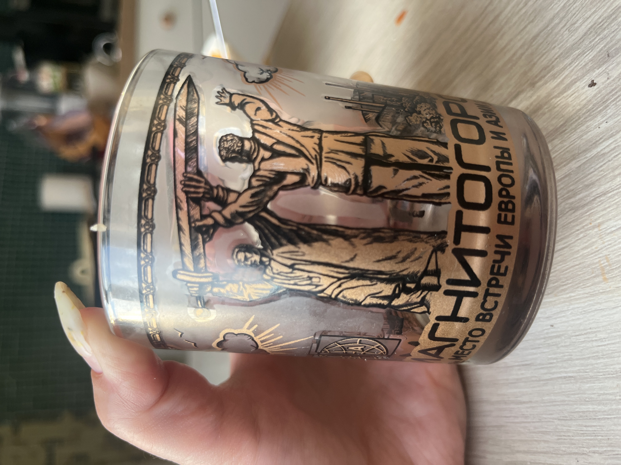 A hand with a painted fingernail is holding a glass cup. The cup has a decorative design featuring two figures holding up a large object, with a sunburst and clouds. Text is visible on the cup in Cyrillic script. The text reads "МАГНИТОГОРСК" and below it "МЕСТО ВСТРЕЧИ ЕВРОПЫ И АЗИИ". This translates to "Magnitogorsk" and "Meeting place of Europe and Asia". Magnitogorsk is a city in Chelyabinsk Oblast, Russia. The background is blurred and appears to be an indoor setting with a countertop and kitchen cabinets.