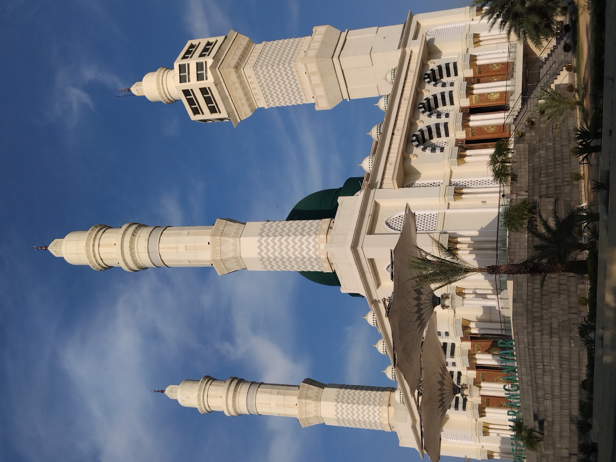 The image shows a grand mosque in Karanganyar, Indonesia, under a bright blue sky with scattered clouds. The mosque features a prominent green dome and three tall, cream-colored minarets, each topped with a crescent moon. The central minaret is the tallest, with a square base and multiple levels, while the two side minarets are cylindrical and slightly shorter. The mosque's facade is adorned with intricate geometric patterns and arched windows. Large, retractable awnings are extended in front of the main entrance, providing shade. The foreground is landscaped with palm trees and other tropical plants, and a stone-tiered platform leads up to the mosque's entrance. Large green letters spelling "PATEN KARANGANYAR" are visible on the stone platform, identifying the location. Several people are visible in the scene, including one person standing on the stairs leading to the entrance and others in the distance. The overall impression is of a majestic and well-maintained religious building bathed in sunlight.