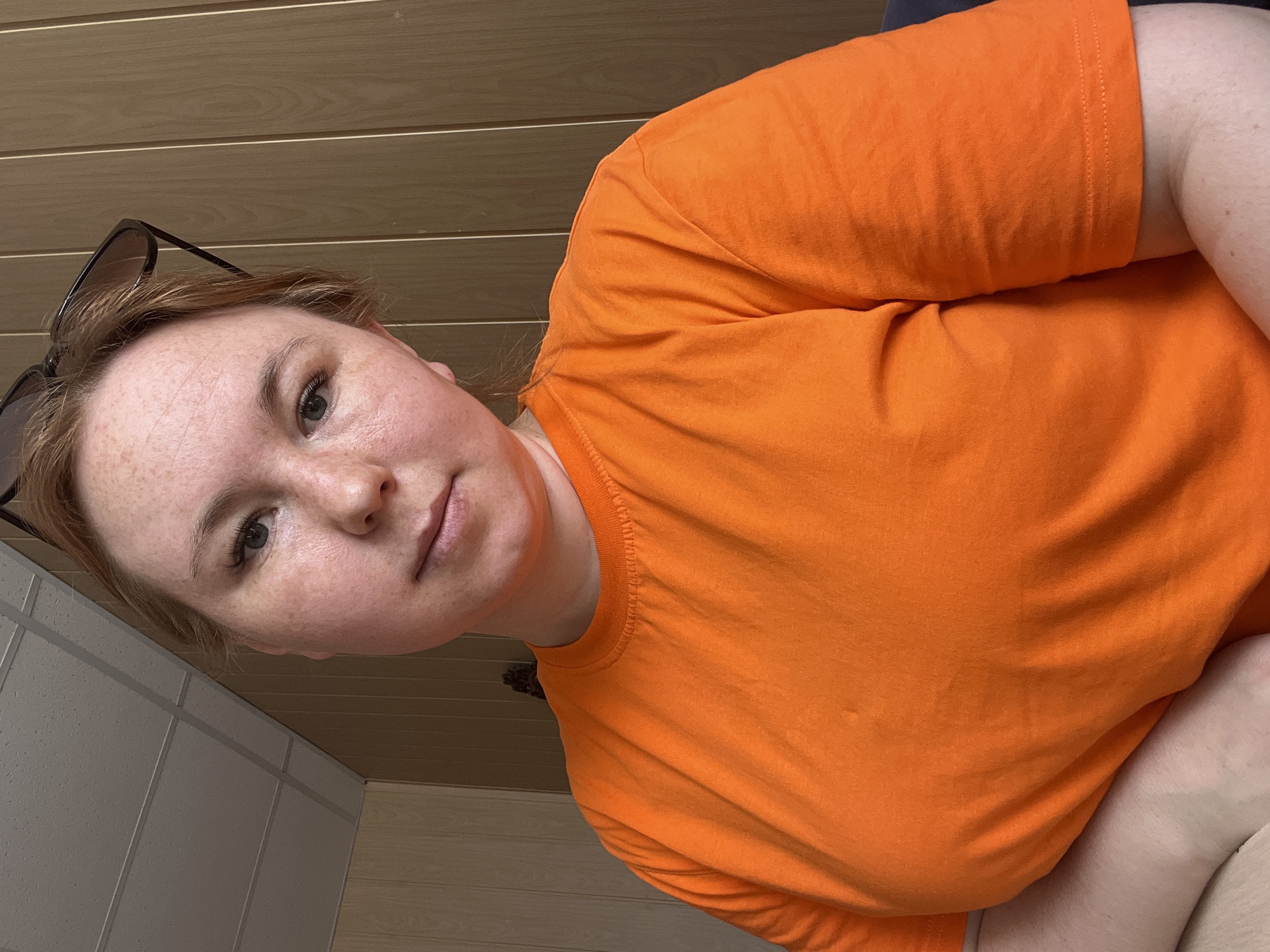 A selfie taken indoors features a fair-skinned woman with freckles, wearing an orange t-shirt. Sunglasses are perched atop her head. The background shows a ceiling with horizontal wooden planks. To the left of the frame, a wall is partially visible with large, light-colored tiles separated by darker seams. There is no information to confirm location in the city of Chaikovsky, Russia.