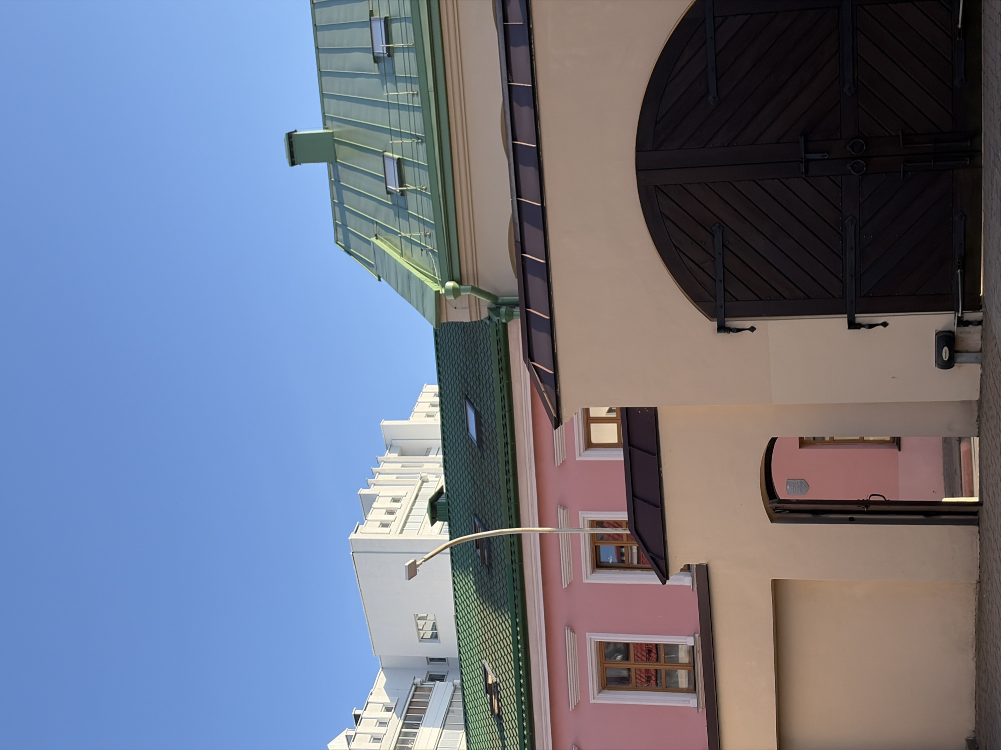 The image depicts a street view in Minsk, Belarus, focusing on the architecture of several buildings. In the foreground, a beige building with a large, arched wooden gate is prominent. The gate is dark brown and features diagonal planks and black metal hardware. To the left of the gate, an open doorway reveals a pink building behind it.
Behind the beige building, a lower pink building with white decorative trim around its windows is visible. The windows have wooden frames and glass panes reflecting the surroundings. Above the pink building, a green roof with several skylights extends across the frame. Further in the background, a modern white building with a geometric facade and numerous windows dominates the skyline. The sky above is a clear, bright blue. A street lamp with a curved pole stands between the pink building and the modern white building. The scene is captured during daylight, indicated by the shadows and the clear sky.