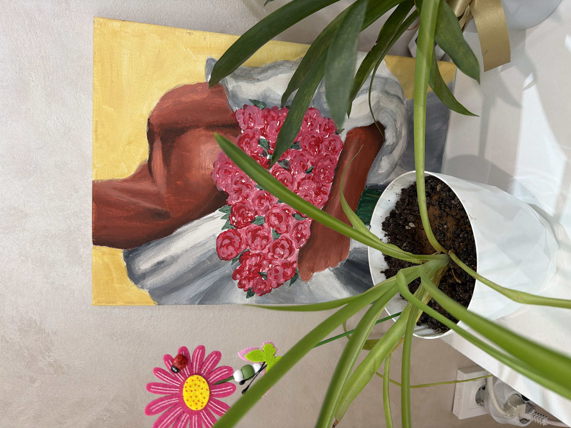A high-angle shot shows a room interior. The scene includes a painting with a brown figure, a white garment, and a bouquet of red roses on a yellow background. The painting is mounted on a light beige wall. A potted green plant with long leaves partially obscures the painting. The plant is in a white pot. A pink artificial daisy with a ladybug and a butterfly adorn the wall below the painting. A white table supports the plant and extends out of frame to the right. Visible on the table are an electrical outlet, a power adapter and associated cables.
There is no information to connect this image to the city of Plovdiv, Bulgaria.