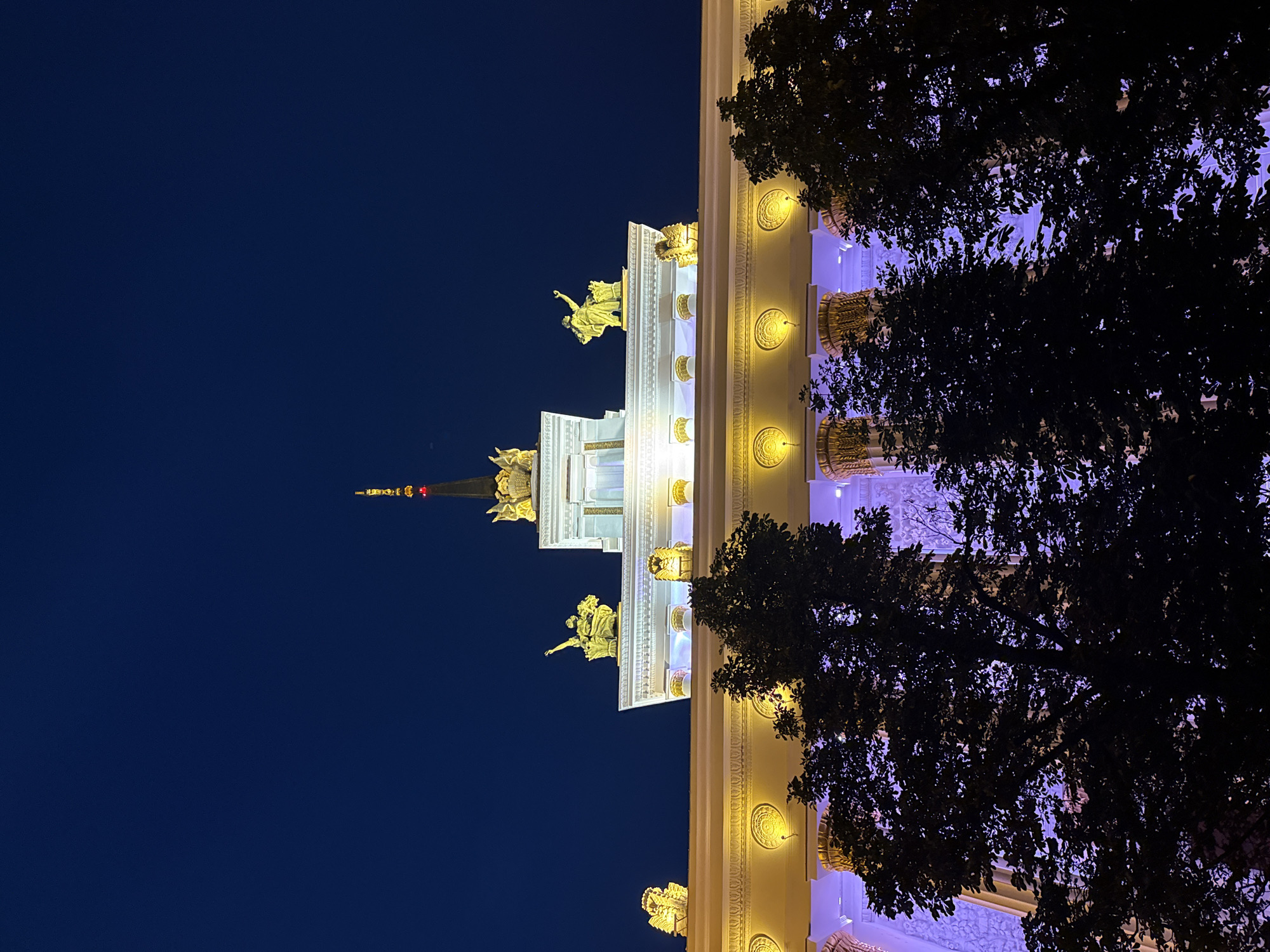 The image shows a low-angle view of the top of a building at night, likely part of the VDNKh (Exhibition of Achievements of National Economy) complex in Moscow, Russia, due to its architectural style featuring classical elements and prominent statues. The building is illuminated with golden lights along its upper facade and with purple lights illuminating the columns and decorative elements below. Silhouetted trees in the foreground partially obscure the view of the building. The sky is a deep, dark blue, indicating it is nighttime. Several golden statues are visible on the roofline, including figures of people and what appear to be lions. The central spire is also illuminated with golden lights and a small red light near its tip. The architectural details suggest a grand and ornate structure, typical of Soviet-era monumental architecture. There are no people visible in the image, and the scene is static.