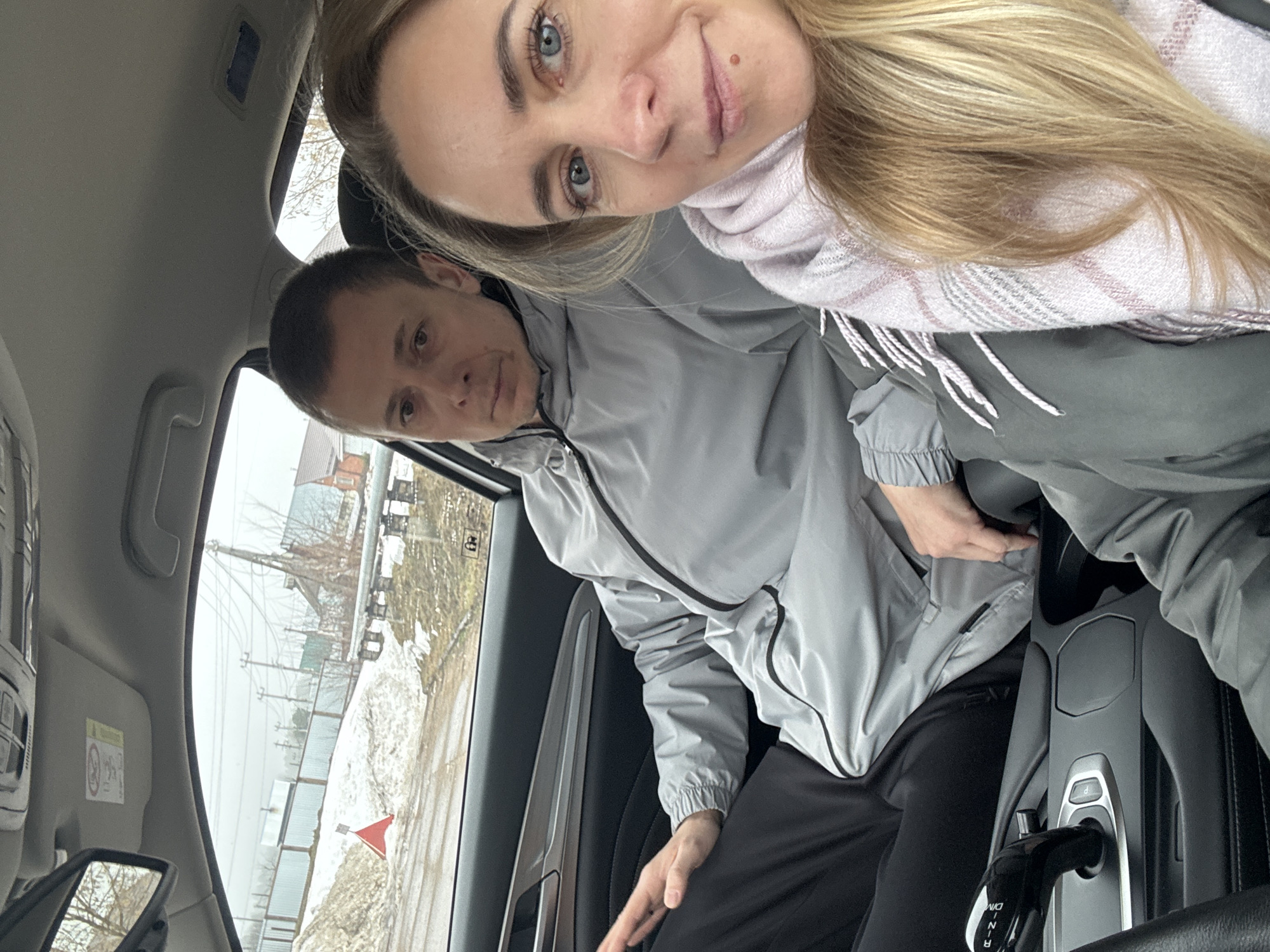 Two individuals are in the front seats of a car. The woman in the passenger seat has blond hair and blue eyes and wears a pink scarf and a gray jacket. The man in the driver's seat has short brown hair and wears a gray jacket. Outside the car window, there is snow on the ground, a red flag, a metal fence, and power lines. In the background are buildings of various colors. The location is Chaikovsky, Russia.