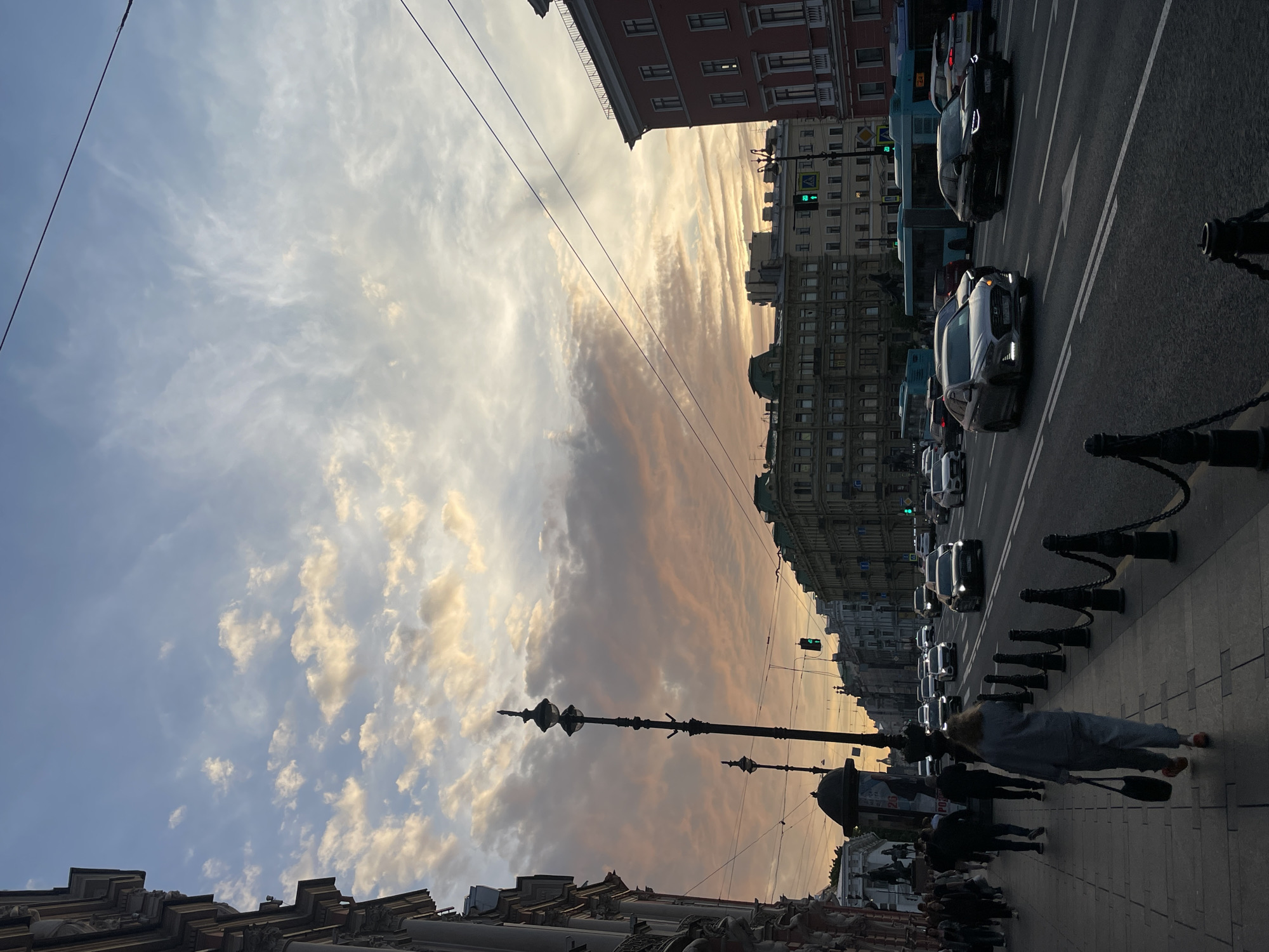 This media file captures a vibrant urban scene, likely a busy street in Saint Petersburg, Russia, at sunset or dusk. The image, originally rotated 90 degrees clockwise, shows a wide street flanked by multi-story buildings and a sidewalk.
In the upper portion of the frame (when correctly oriented), the sky is a striking feature, filled with clouds illuminated by the low-angled sun. Golden, orange, and light brown hues stretch across the horizon, blending into lighter blue and white clouds further up. Overhead wires cut across the sky.
The street below is busy with traffic. Several cars, including modern sedans and what appear to be buses, are visible, many with their headlights on, indicating dimming light. Traffic signals are present, with at least one green light visible. On the right side of the street, a sidewalk is bustling with pedestrians. In the foreground, a person with reddish hair, wearing a gray sweatshirt and pants, and carrying a black bag, walks along the sidewalk, her back to the viewer. Other pedestrians are visible further down the sidewalk.
Ornate, black streetlights and bollards with chains line the sidewalk, adding to the classic European city aesthetic. The buildings lining the street are grand and varied in architectural style, some appearing older with intricate details, others more contemporary. One structure in the foreground appears to be a monument or part of a building facade with sculptural elements. Visible text on a kiosk or sign near one of the streetlights is present but indistinct due to distance and resolution. The overall atmosphere suggests the end of a day, with soft, warm light casting long shadows and illuminating the clouds dramatically.