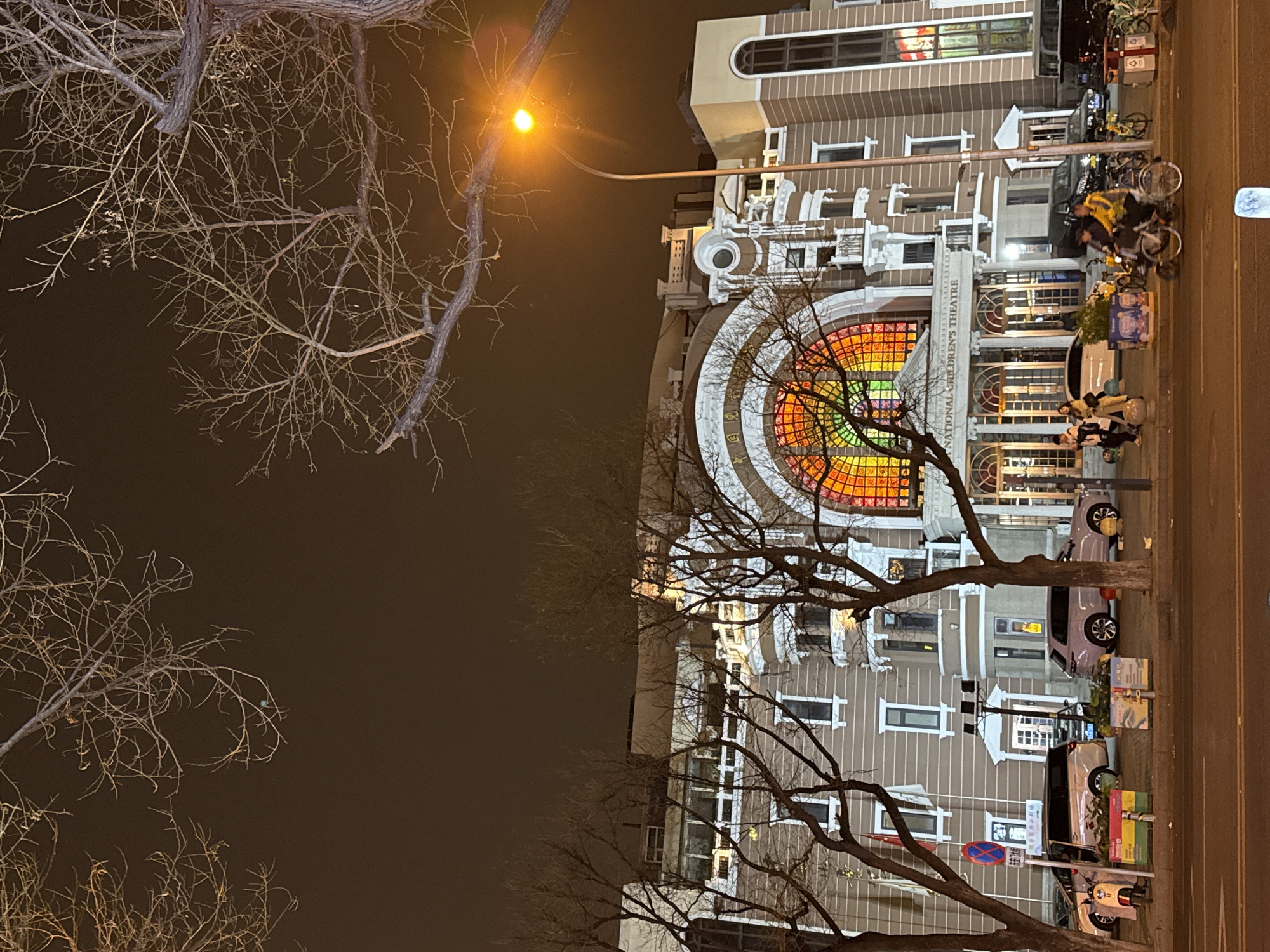 The image depicts the National Children's Theatre in Dongcheng District, China, at night. The building has a large, colorful arched window above the entrance. People and bicycles are present on the sidewalk. Bare trees frame the building. A street light illuminates the scene. Cars are parked along the street. The sky is dark.