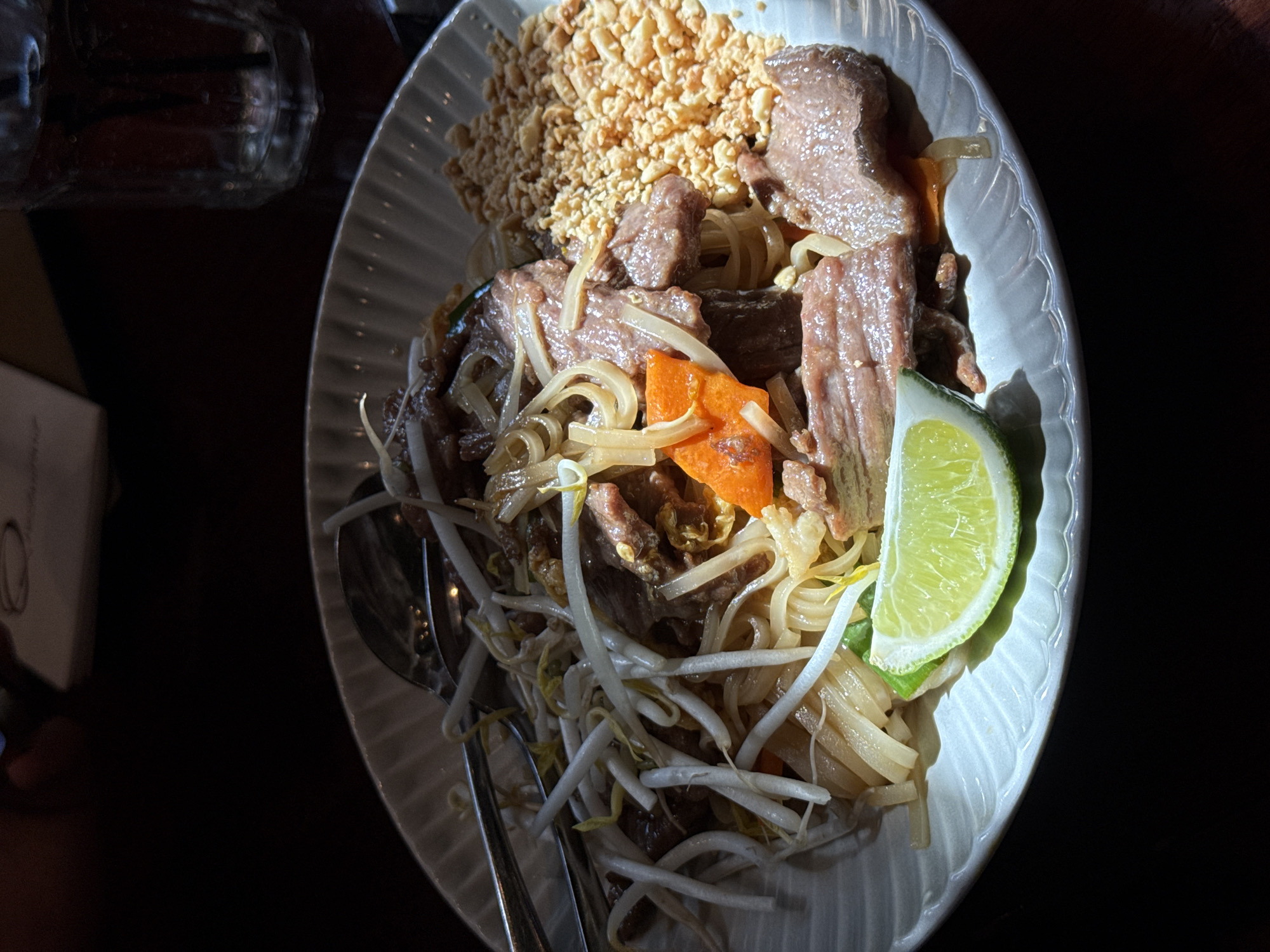 The image shows a plate of Pad See Ew, a stir-fried noodle dish, likely served in a restaurant. The dish contains wide rice noodles, beef, bean sprouts, carrots, crushed peanuts, and a wedge of lime. Two metal spoons are resting on the plate. A glass and a menu are partially visible in the background. The lighting suggests an indoor setting with a light source coming from the right side. The location is unknown, but the prompt requests the city of Boston, United States of America.
