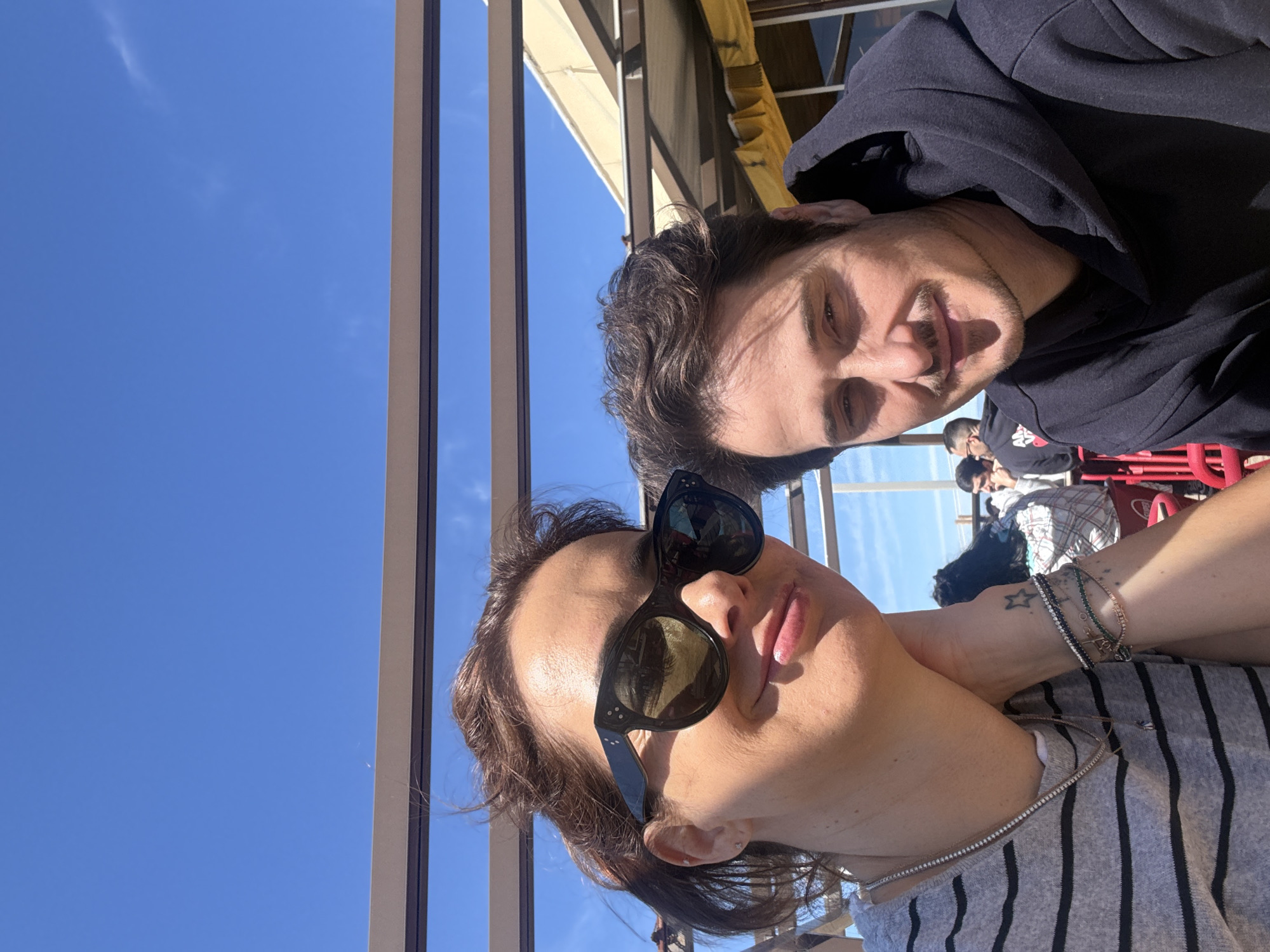 Two people are pictured in a selfie taken outdoors on a sunny day. The woman in the foreground wears sunglasses and a striped shirt. The man is behind her and slightly to the right, wearing a dark hoodie. In the background, other people are seated at tables. The clear blue sky is visible above a pergola-like structure. Red seating and other diners can be seen. The setting appears to be an outdoor dining area. Although a specific location in Vila Nova de Gaia is unknown, the architectural details and outdoor dining environment suggest a riverside restaurant or cafe.