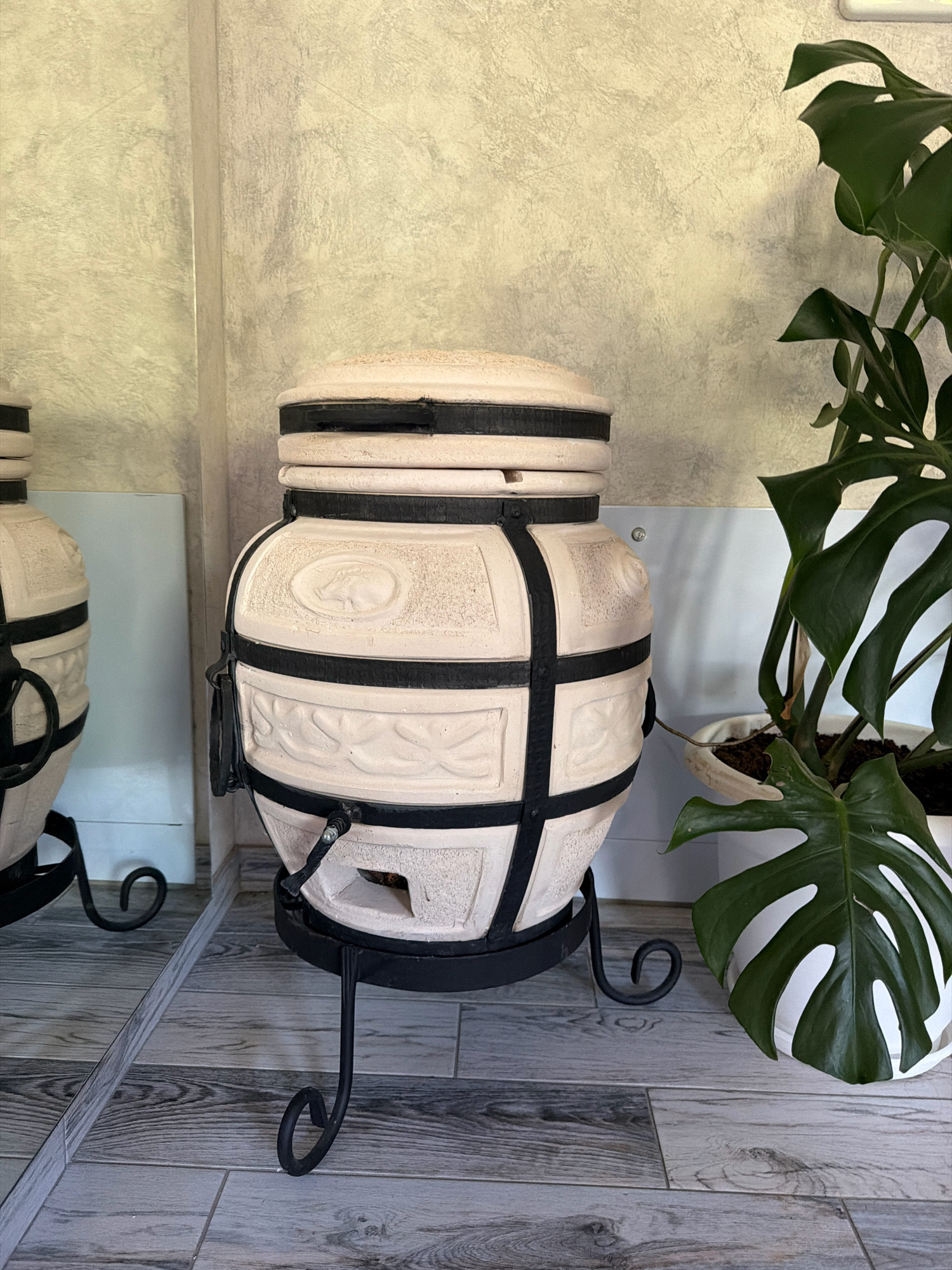 A tandoor oven, made of light-colored ceramic and reinforced with black metal bands, stands on a black metal stand with curved legs. The oven has intricate decorative embossing and a small opening at the bottom. To the right of the tandoor, a large potted Monstera plant with broad, fenestrated leaves is visible. The floor is covered in light gray wood-look tiles. A large mirror on the left reflects the tandoor and the surrounding environment, including another similar tandoor oven partially visible. The wall behind the objects has a textured, pale gray finish. The scene appears to be indoors, likely in a home or a commercial space, given the presence of decor and flooring. No people are present in the image. The setting is identified as within the boundaries of Перовское сельское поселение, Ukraine.