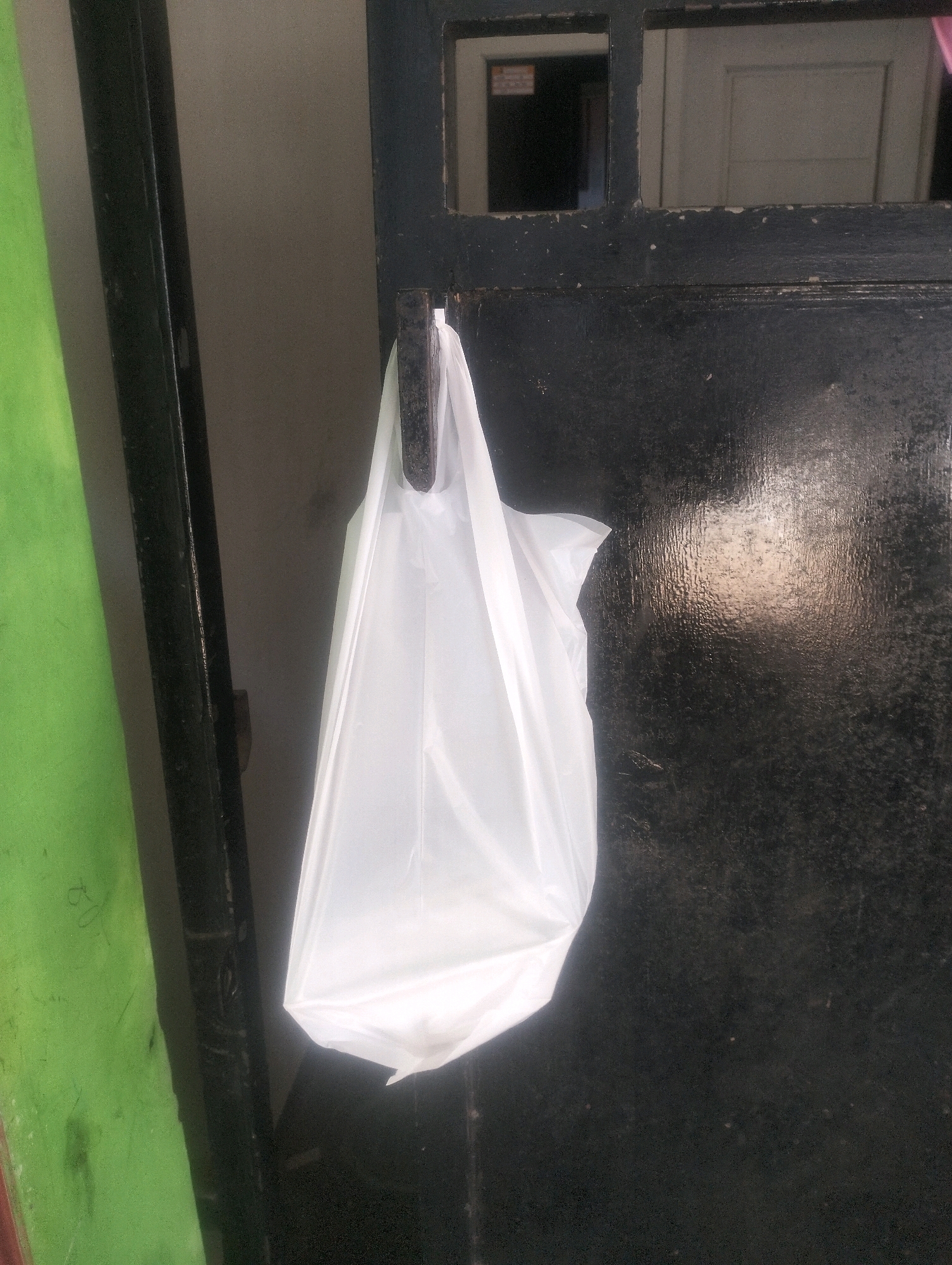 A white plastic bag hangs from a black metal hook on a black door. The door has a small square window with a grid pattern in the upper section, revealing a glimpse of a hallway or room inside. To the left of the door, a portion of a bright green object, possibly a wall or a large piece of fabric, is visible. The environment appears to be outdoors or in a transitional space between indoor and outdoor areas. The lighting suggests it is daytime. This scene is located in Purwokerto, Indonesia.