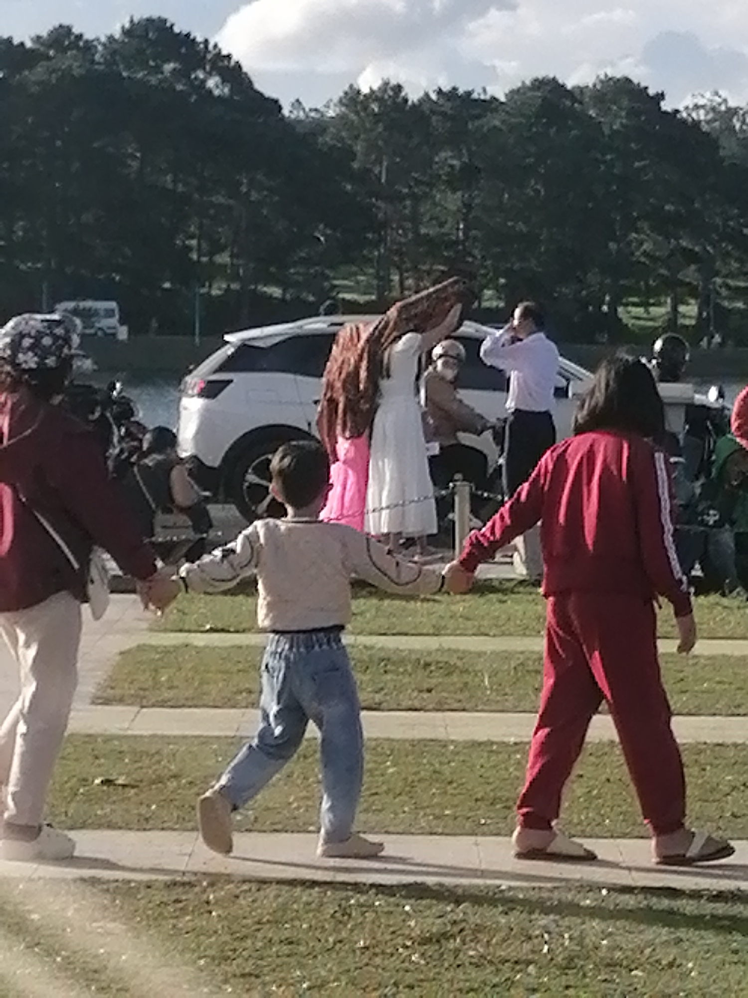 This media file depicts a scene with several people in an outdoor setting, possibly a park or a public gathering space, in Da Lat, Vietnam. The time of day appears to be daytime, with bright sunlight casting shadows, suggesting a sunny or partly cloudy weather condition.
The primary subjects are a group of individuals of varying ages. In the foreground, a young child in a light-colored long-sleeved shirt and blue jeans is holding hands with two other people. To the child's left, a person in a maroon jacket and light-colored pants is visible from the waist down, also holding the child's hand. To the child's right, a person in a maroon tracksuit is walking. The clothing suggests a casual setting.
In the mid-ground, there are other people and objects. A white SUV is parked behind the individuals in the foreground. Further back, there are trees, a lush green landscape, and a cloudy sky. There are also hints of other vehicles, including what might be a van in the far left background. Some people appear to be engaged in conversation or observing the surroundings. One person in the background is wearing a light-colored shirt and dark pants and appears to be adjusting their clothing or hair. Another individual is wearing a white dress, and someone else is in a brown outfit.
The overall mood seems relaxed and casual, with people enjoying an outing. The presence of a paved pathway suggests a well-maintained public area. The dense trees in the background are characteristic of mountainous or forested regions, which aligns with the known geography of Da Lat. No visible text is present in the image.