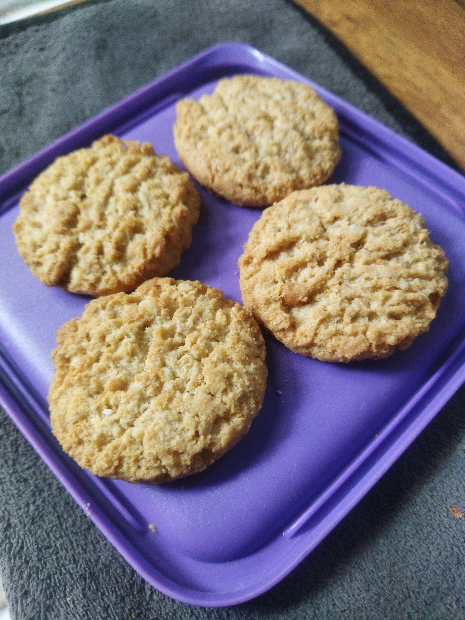 Four golden-brown cookies are arranged on a purple plastic tray. The cookies are round and have a slightly uneven, textured surface, suggesting they might be homemade or have an oat-based ingredient. They are positioned in a loose cluster on the tray, with two cookies in the foreground and two slightly behind them. The tray itself is angled, and its surface shows some light reflections.
The tray is placed on a dark grey, textured surface, likely a cloth or fabric. To the right of the tray, a portion of a wooden surface is visible, indicating a table or countertop. The background is blurred, suggesting a shallow depth of field.
The scene is a close-up shot, focusing primarily on the cookies and the tray. The lighting appears to be natural, casting soft shadows and highlighting the textures of the cookies.
There are no people visible in the image, nor any explicit indication of the city of Tarai Bangun, Indonesia. The description is limited to the visual elements present in the photograph.