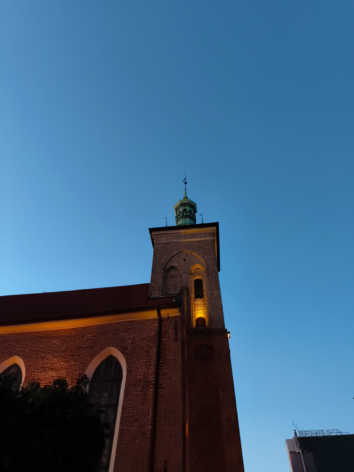 The photograph captures a twilight scene of a red brick building, likely a church, with a prominent tower. The building features arched windows and a red tiled roof. The tower has a green, octagonal spire topped with a small flag. Warm light emanates from some of the tower's windows and along the edge of the roof. In the background, the clear blue sky suggests early evening. To the right, a sign reading "Mercure HOTEL" is visible on another building, indicating an urban setting. The presence of the hotel sign strongly suggests the location is in or near a hotel in Gdańsk, Poland. No people are visible in the frame.