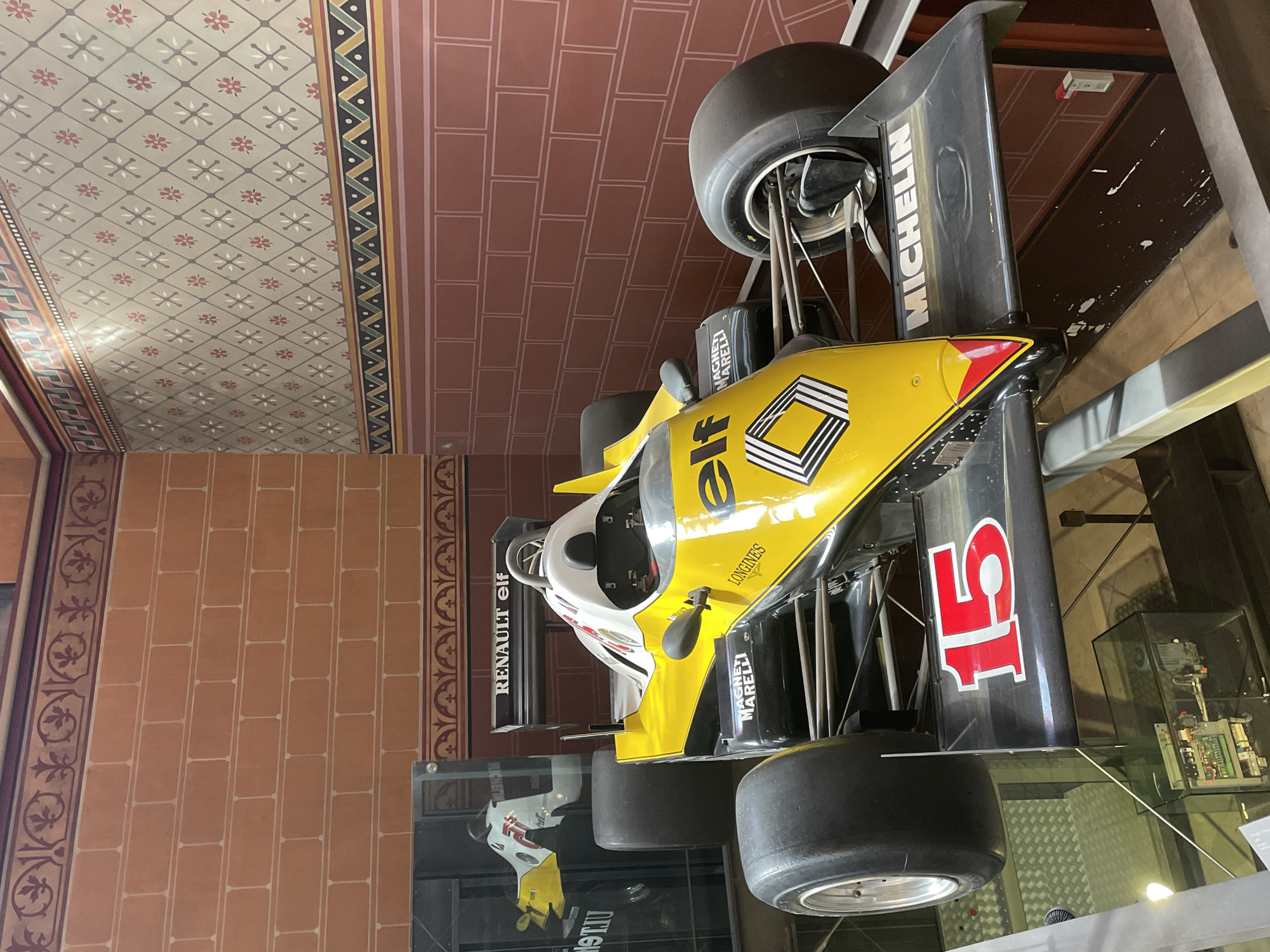 The image depicts a vintage Formula 1 racing car, predominantly yellow with black and white accents, displayed indoors. The car features prominent sponsor logos such as "RENAULT elf," "MAGNETI MARELLI," "LONGINES," and "MICHELIN." The number "15" is visible in red on the rear wing. The car is positioned on a raised platform, possibly in a museum or exhibition space. The background consists of walls adorned with decorative brick patterns and intricate geometric wallpaper. A reflection of a similar racing car is visible on a glass surface to the left of the frame. The lighting suggests an indoor environment, with some light reflecting off the polished surfaces of the car. The scene is static, with no people or activity. While the image is taken in Paris, France, the specific location within the city is not discernible from the visual information provided.